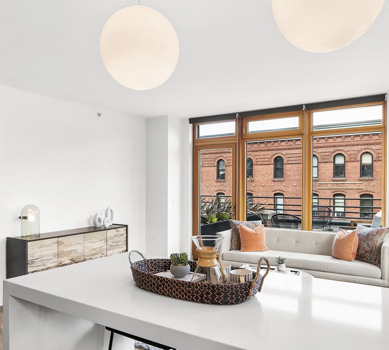 Modern living room with white walls, wooden window frames, a cream sofa with orange and patterned pillows, a balcony with outdoor furniture, and decorations on a sideboard.