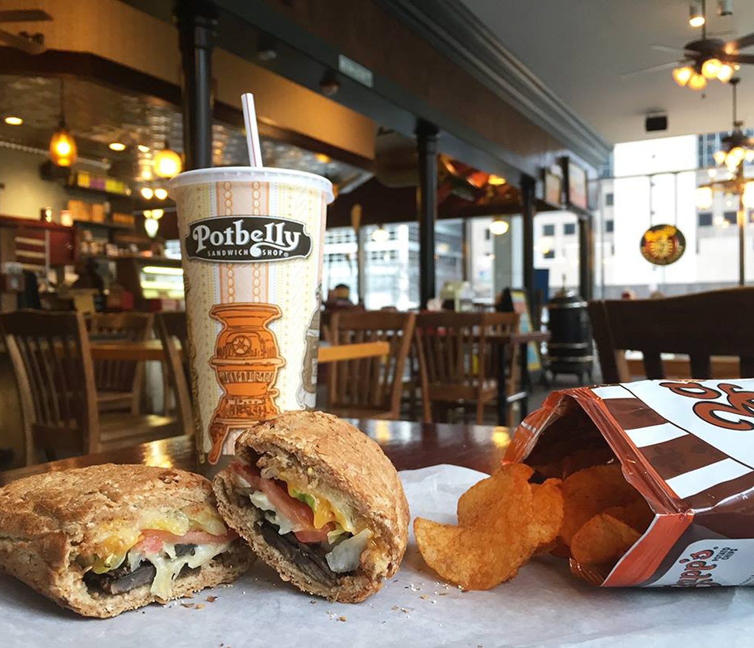 A sandwich wrap, potato chips, and a drink on a table inside Potbelly Sandwich Shop with empty chairs and windows in the background.