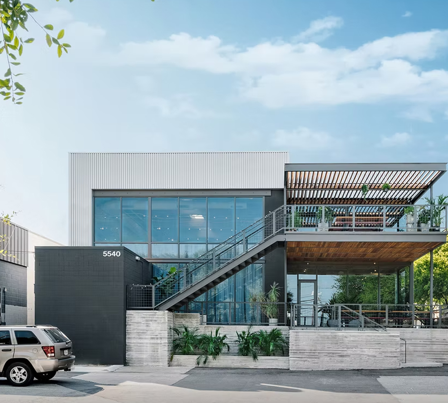 Modern commercial building with large glass windows, metal siding, a rooftop patio with plants, an external staircase, and a parking lot in the foreground.