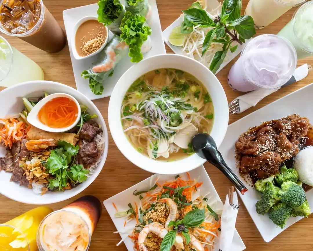 An assortment of Thai dishes on a wooden table, including soup, salad, fried chicken with broccoli, and drinks like iced coffee and colorful beverages.