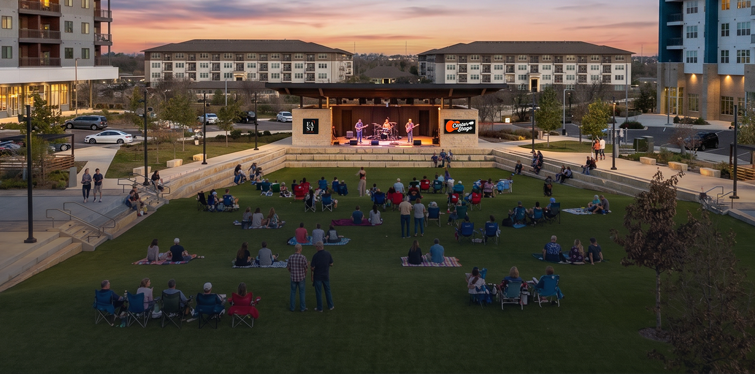 An outdoor concert at dusk with a small band on stage and a seated audience on grass and benches in a park surrounded by modern residential buildings.