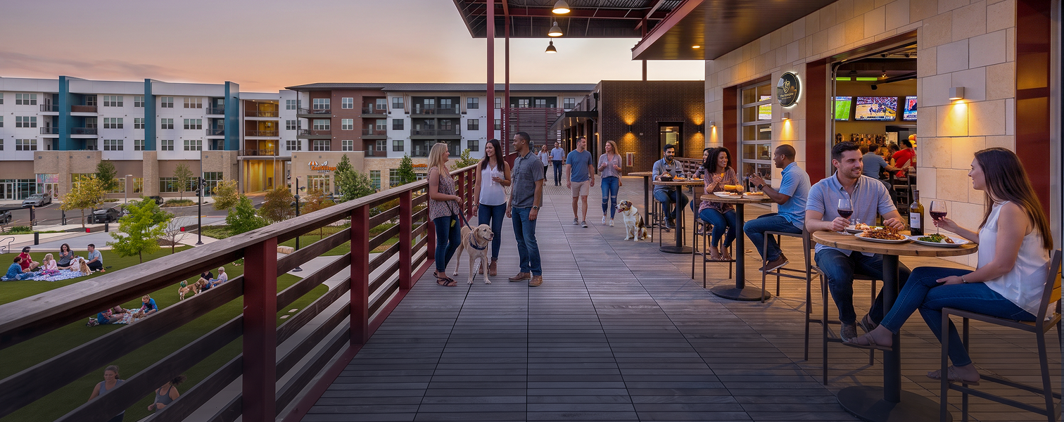 People dining and socializing on a rooftop patio at sunset, with apartment buildings and a park in the background.