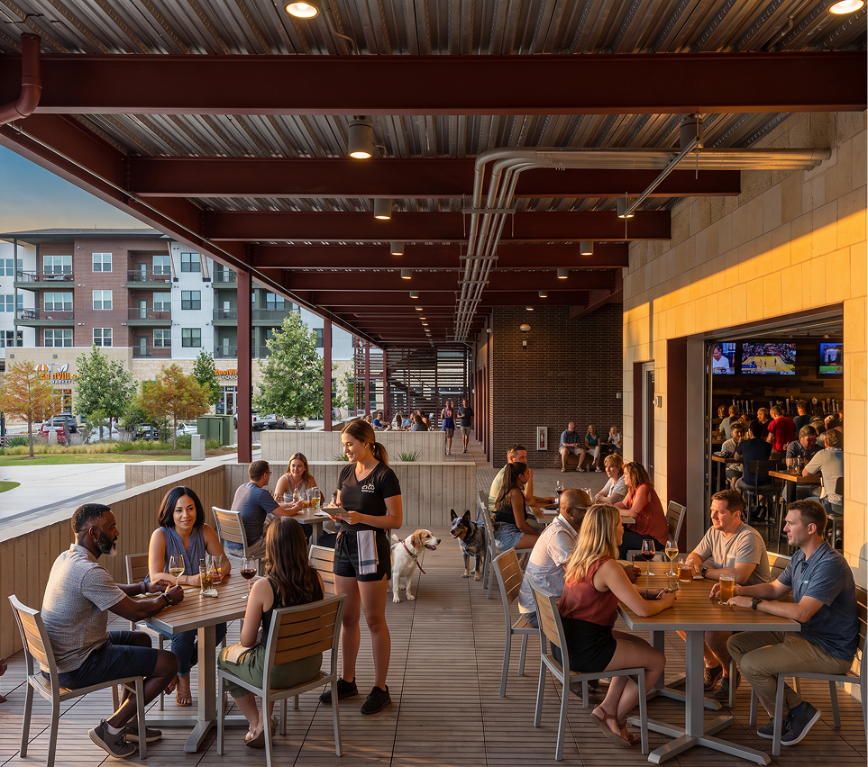 People dining on an outdoor patio at a restaurant at East Village with some dogs and a view of apartment buildings in the background.