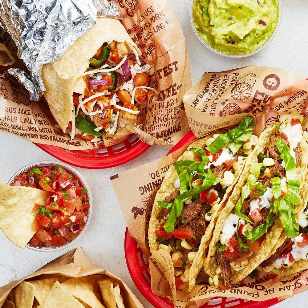 Two tacos with shredded meat, lettuce, tomatoes, and other toppings in red baskets. A side of pico de gallo and guacamole, and a basket of chips.
