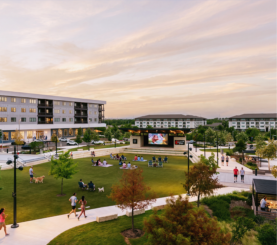 East Village during sunset with people sitting on the grass and walking, an outdoor movie screen showing images, surrounded by trees, apartment buildings, and parking lot.