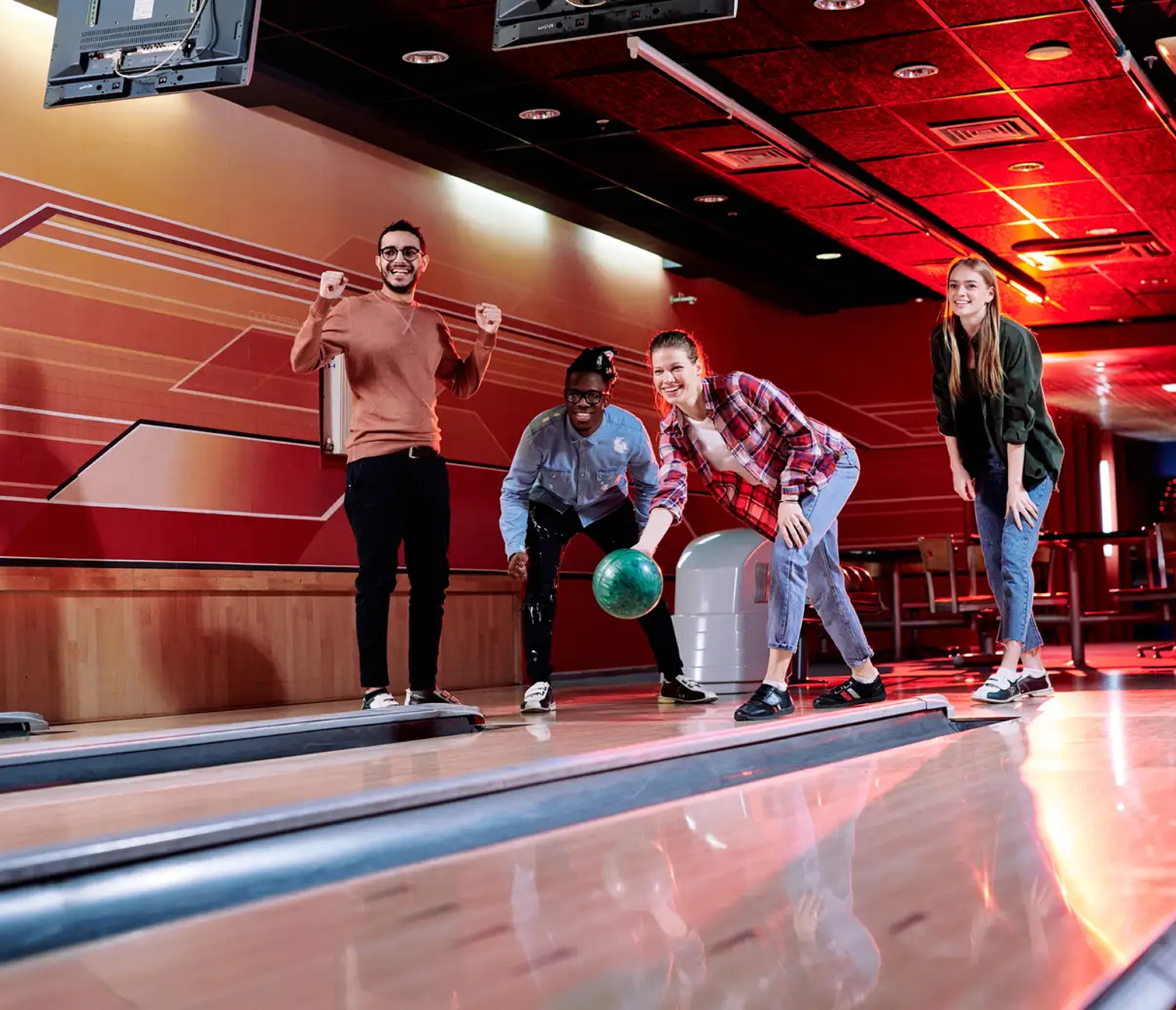 Group of four friends at a bowling alley, one is about to bowl, and others are cheering, with red lighting and wooden lanes.