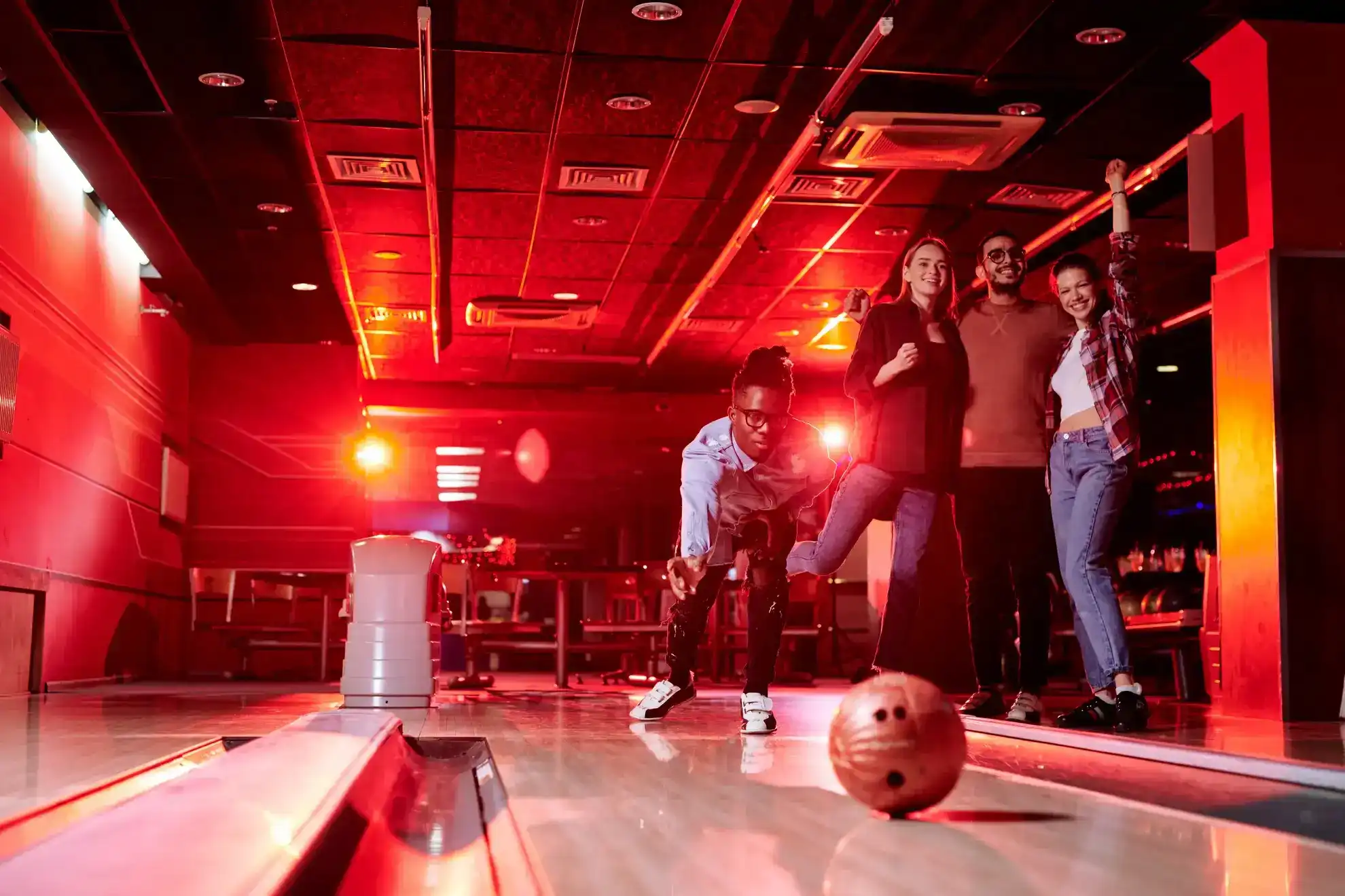 Three young friends, two women and one man, joyfully pose and celebrate in a bowling alley, with one person preparing to bowl and the others smiling, surrounded by vibrant red lighting.