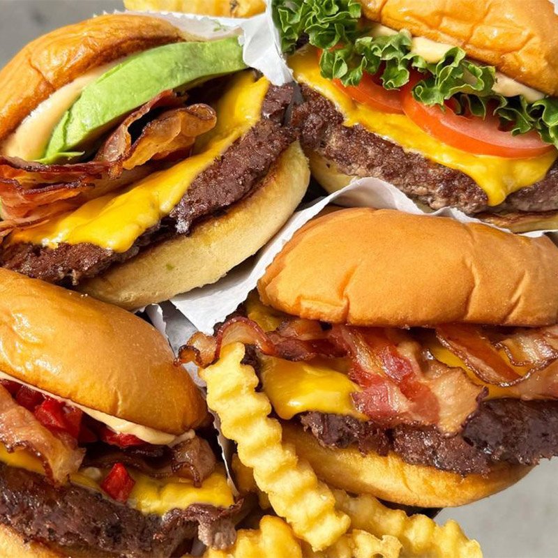 Close-up of three cheeseburgers with crispy fries on top, with bacon, lettuce, tomato, and cheese visible in the burgers.