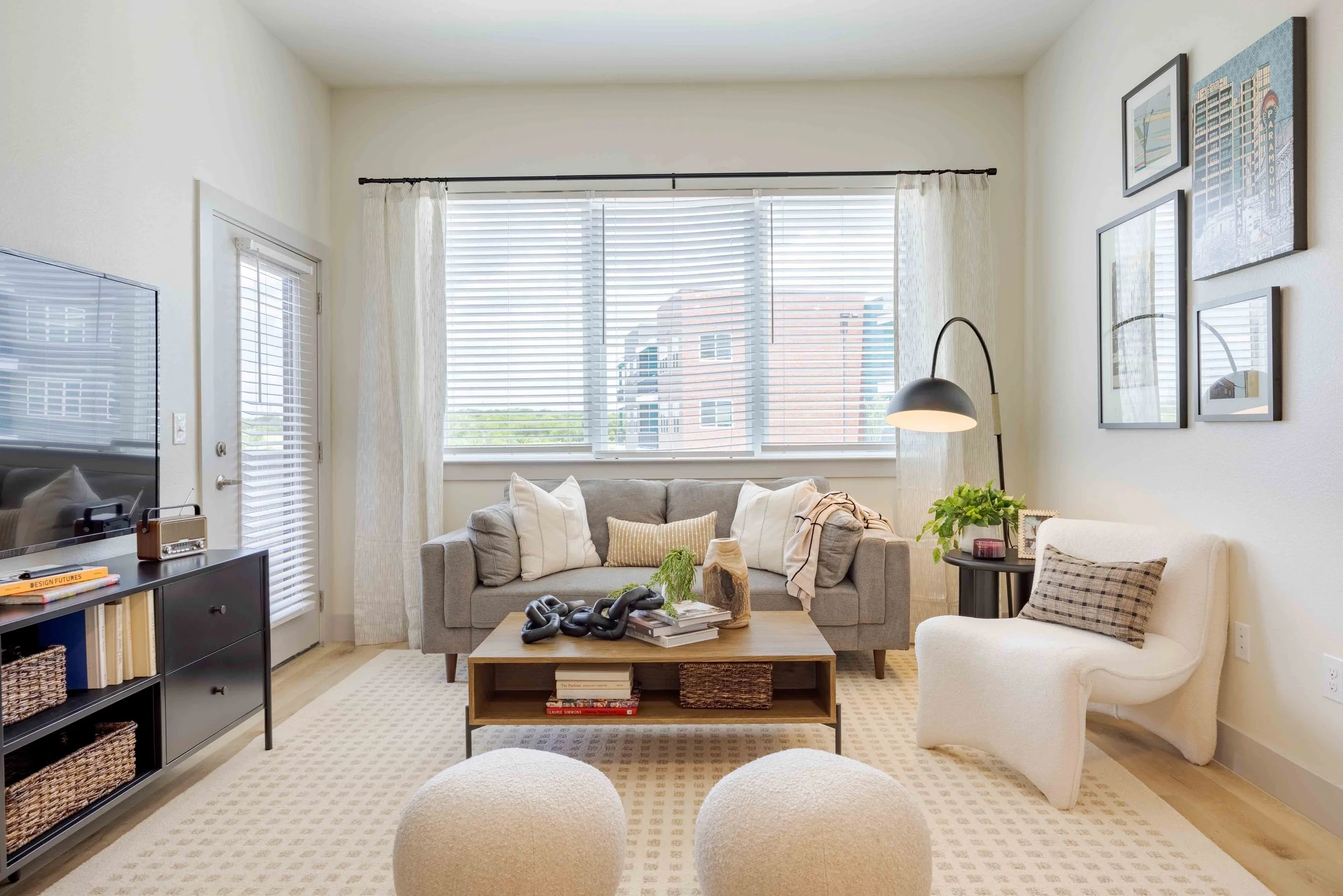 Living room with beige walls, large window with blinds, gray sofa with pillows, side chairs, wooden coffee table, black TV stand with books and decorations, wall art, floor lamp, potted plant, beige patterned rug.