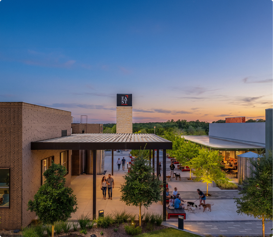 People walking and sitting at an outdoor shopping or dining area during sunset, with trees and modern buildings.