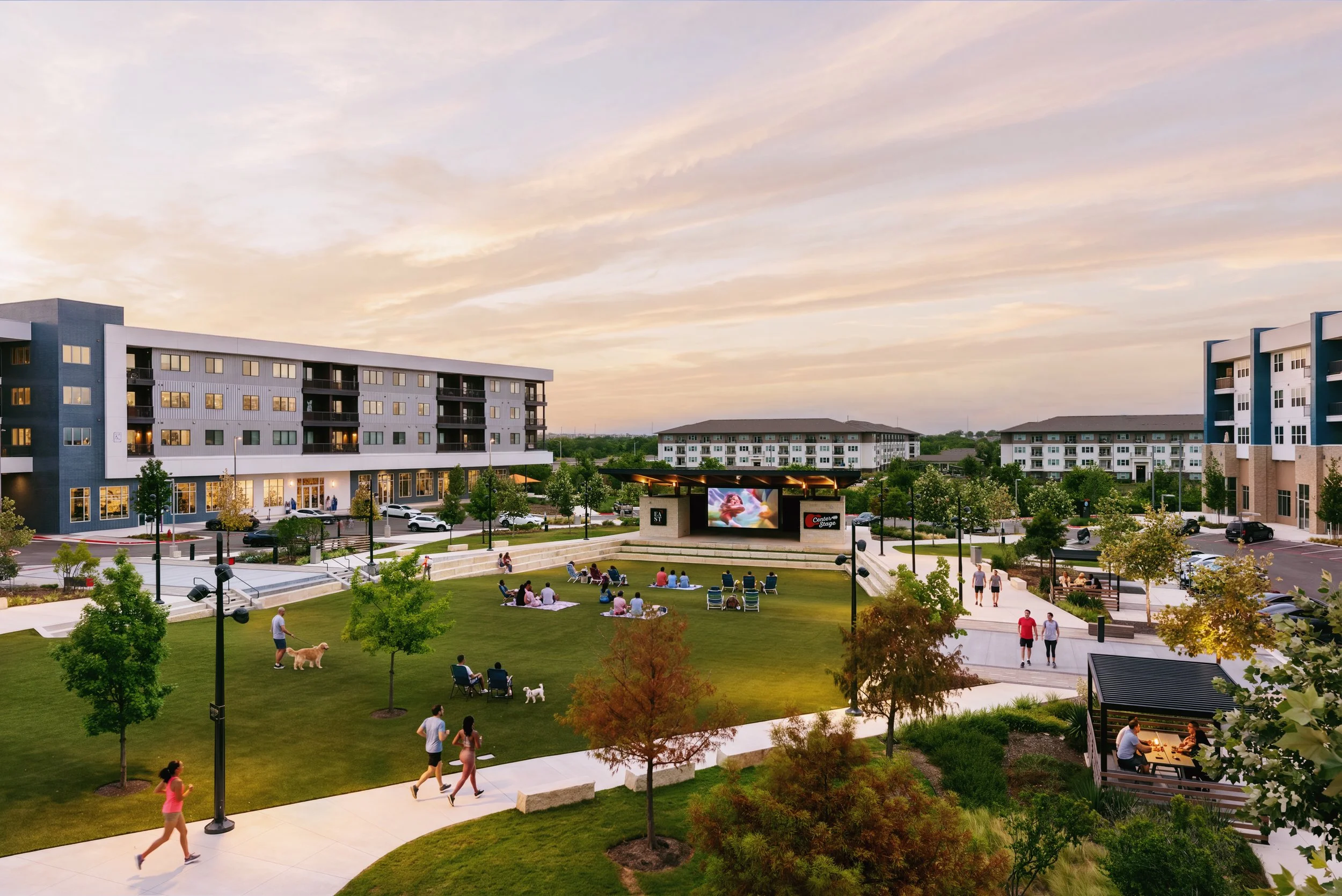 A modern apartment complex with multiple buildings surrounding a landscaped courtyard. The courtyard features a grassy area with people sitting and walking, some with dogs, and a small outdoor stage with a screen. The sky is partly cloudy with a soft evening light.