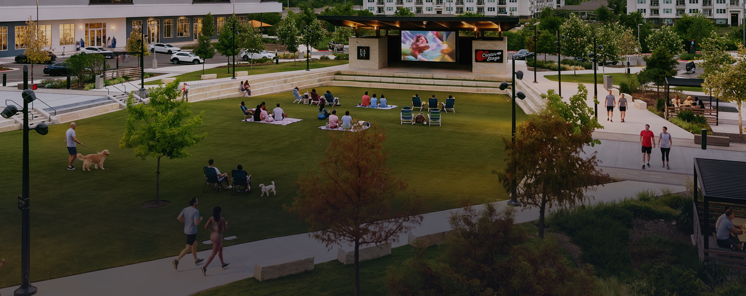 An outdoor amphitheater with a grassy area, trees, and a stage with a large screen, where people are sitting on blankets and chairs watching a movie. Walkers, dogs, and people strolling nearby.