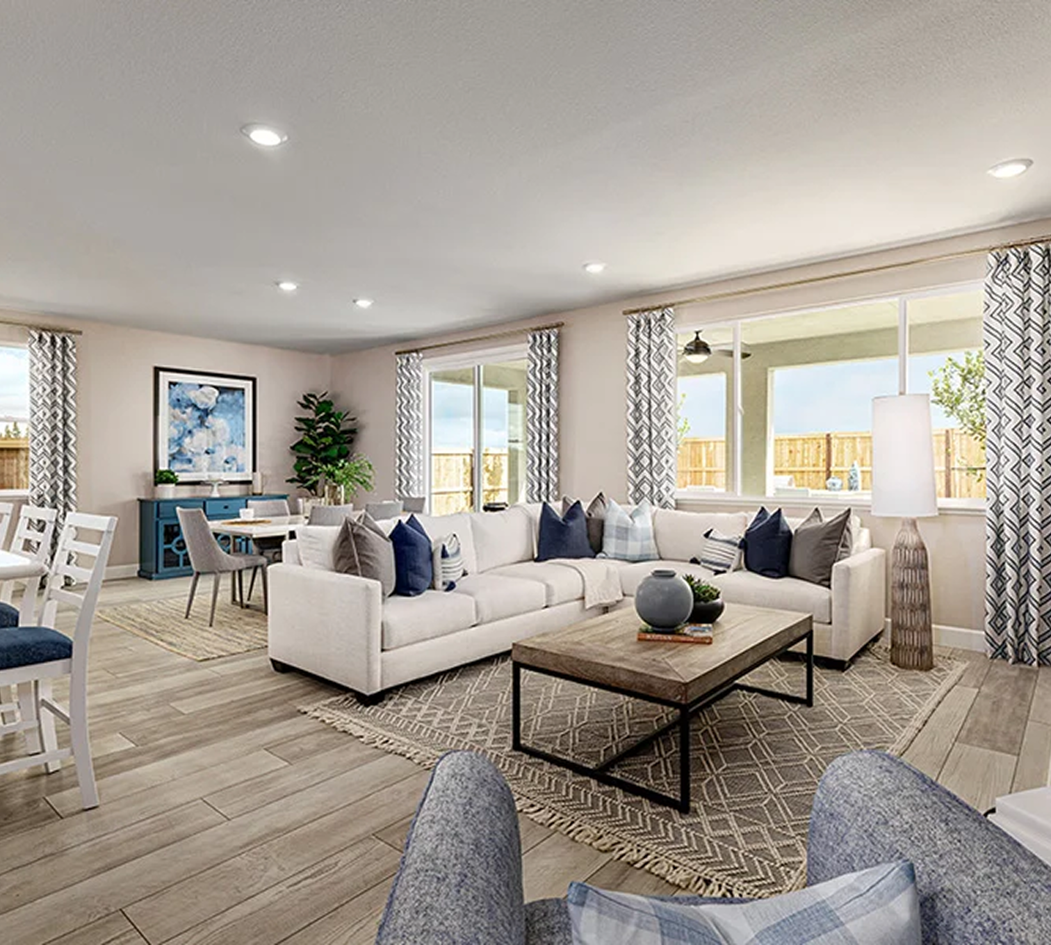 Living room with white sofa, blue and gray pillows, wooden coffee table with vases, patterned area rugs, large windows with patterned curtains, and a ceiling with recessed lighting.