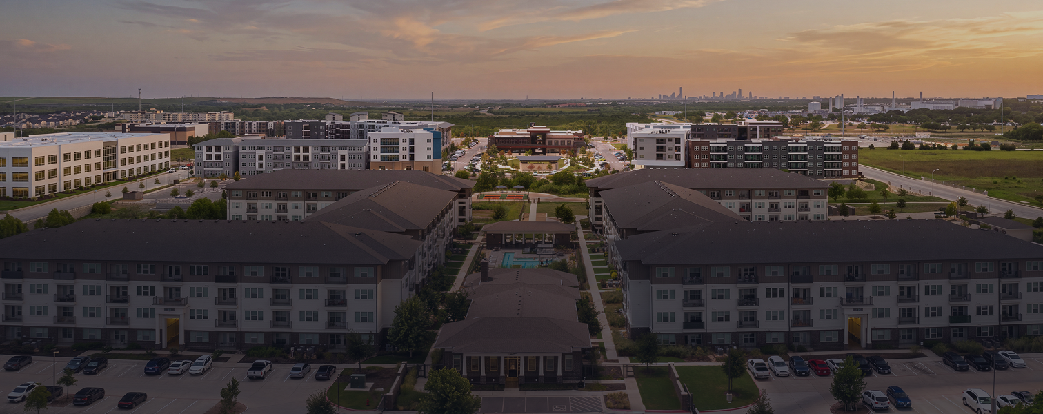 Aerial view of a modern apartment complex at sunset with parking lots, green spaces, and a city skyline in the distance.