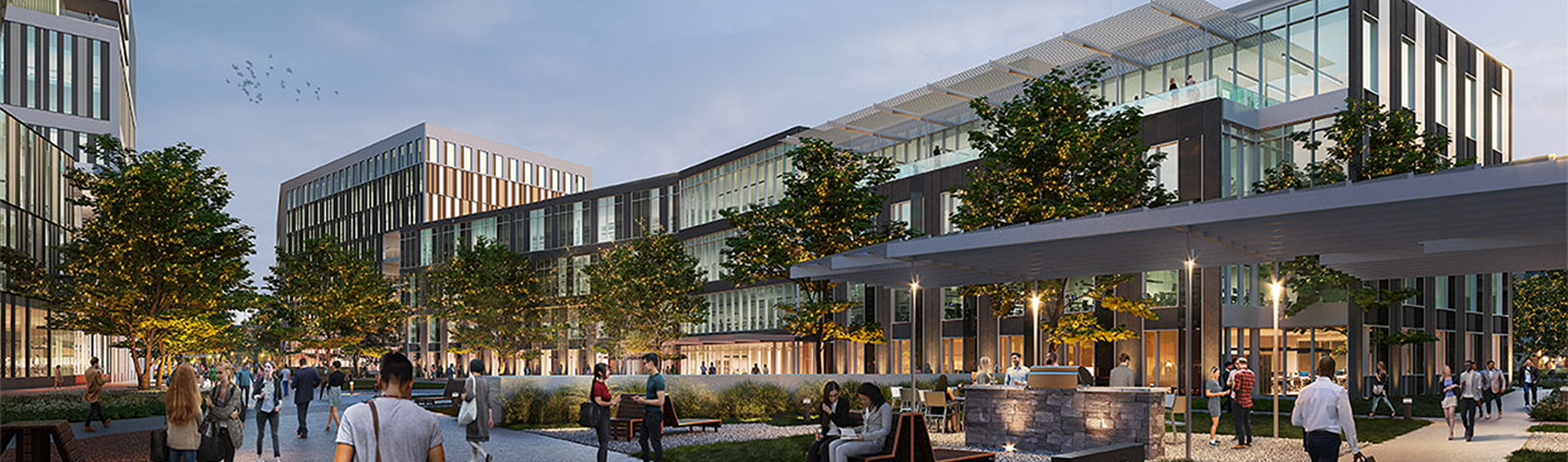 A modern urban outdoor area with people walking, sitting on benches, and socializing in front of commercial buildings with glass facades and trees, during early evening.