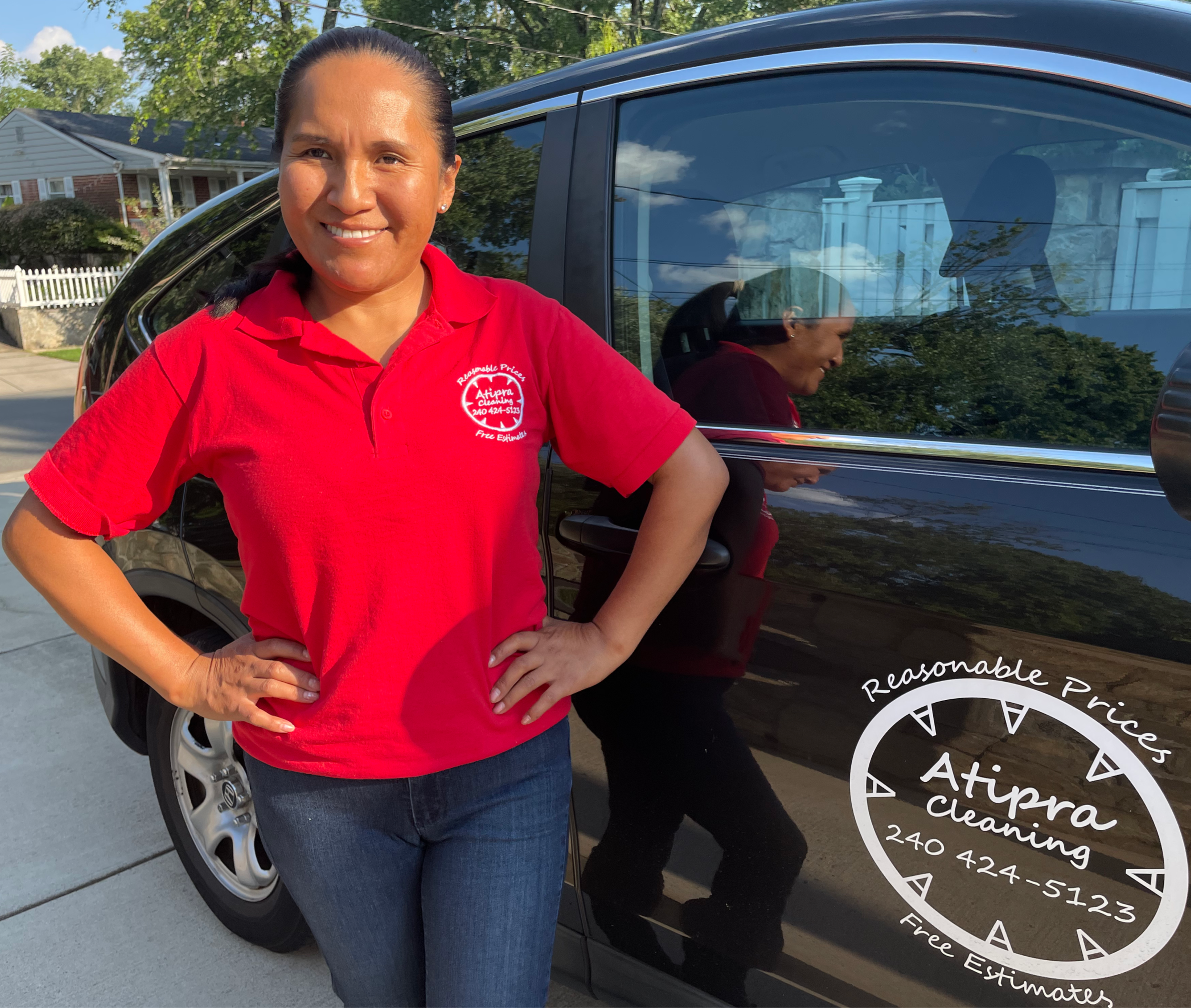 A woman with dark hair tied back, wearing a red polo shirt with a company logo, standing outside next to a black vehicle that has a circular vinyl decal promoting a cleaning business. She is smiling and has her hands on her hips in front of a residential neighborhood with trees and houses.