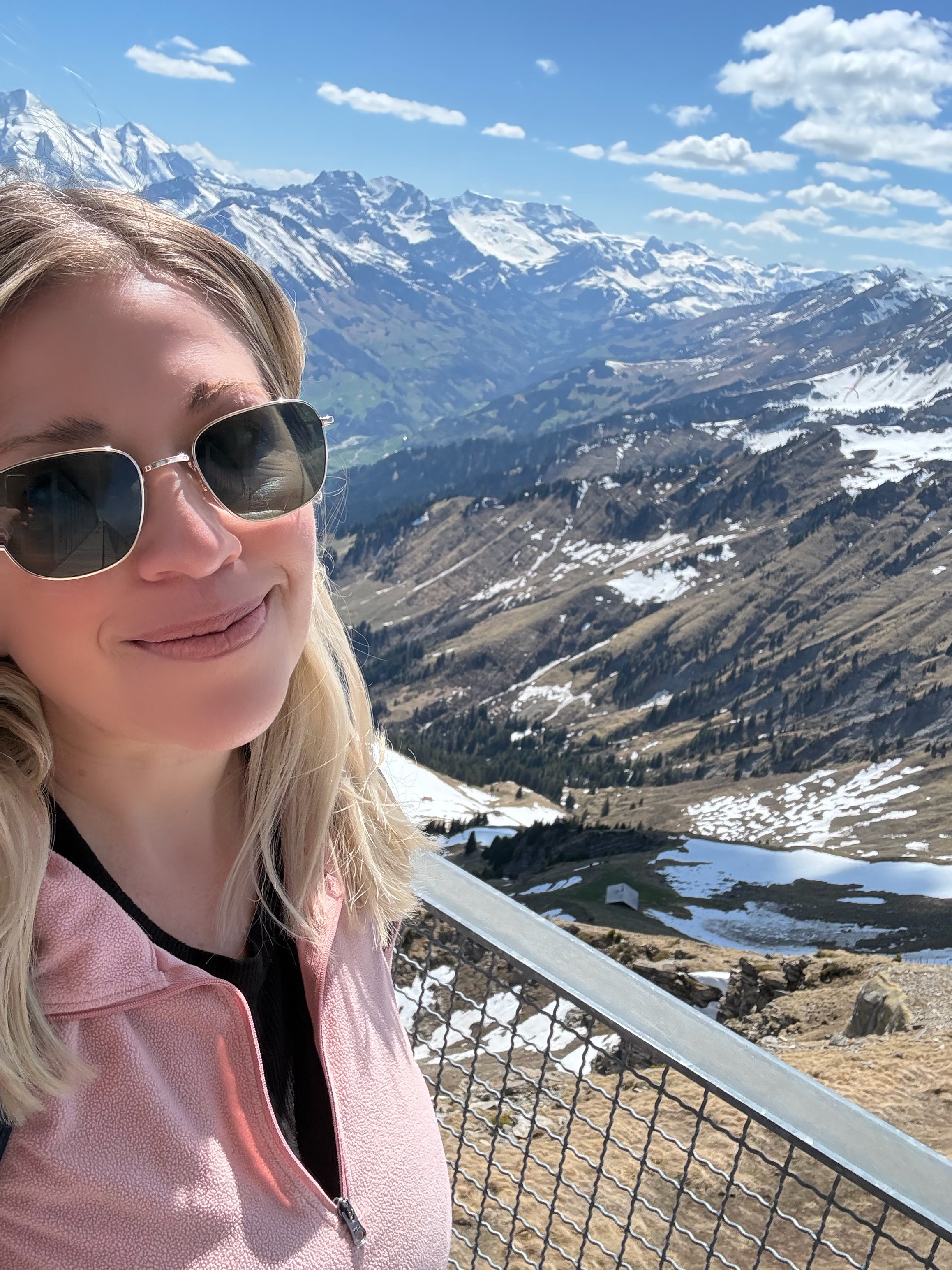 A woman with blonde hair, wearing sunglasses and a pink jacket, standing on a mountain overlook with snow-capped mountains and a partly cloudy sky in the background.