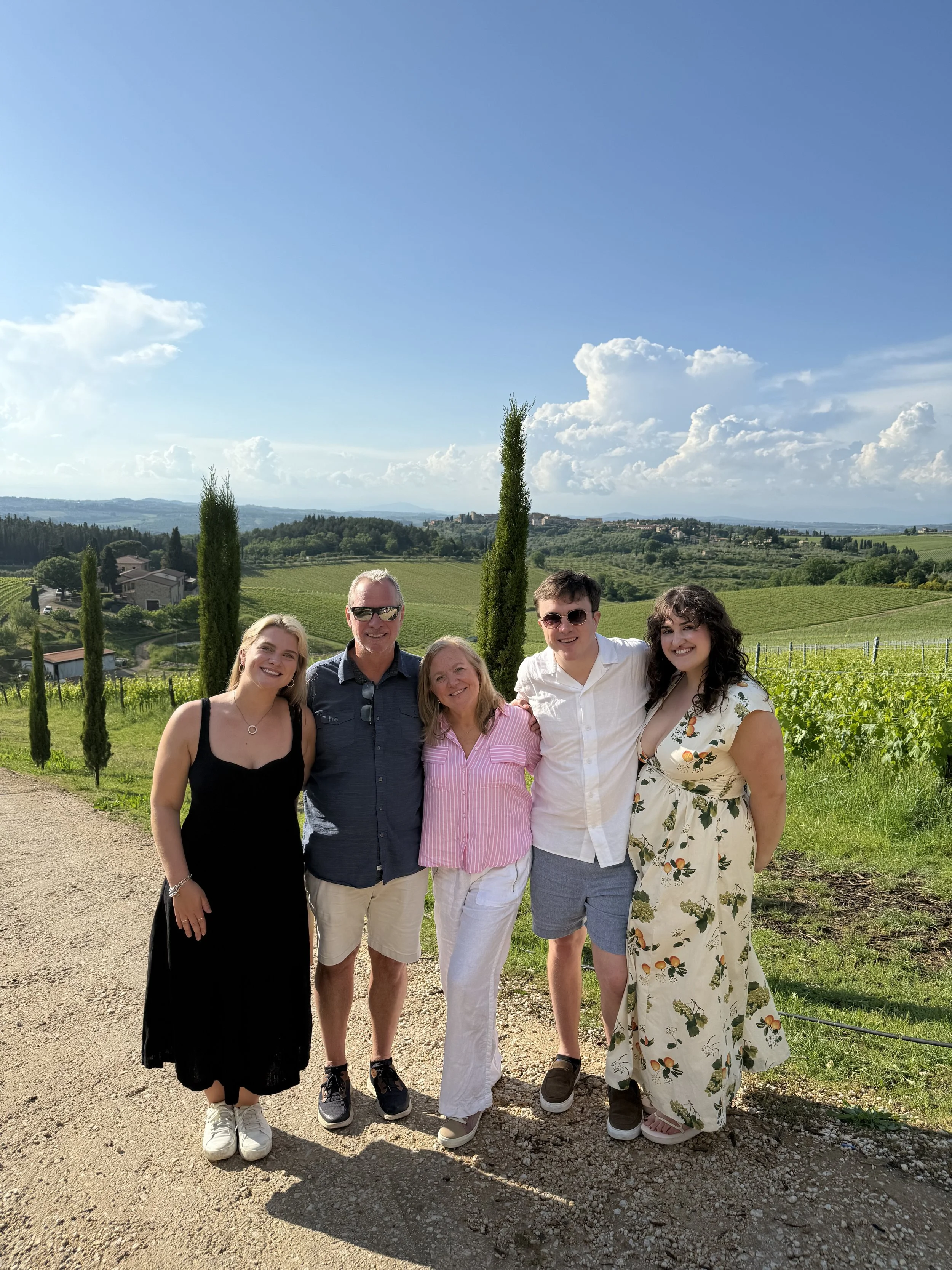 Family photo in a scenic landscape with rolling hills, vineyards, and cypress trees under a partly cloudy blue sky.