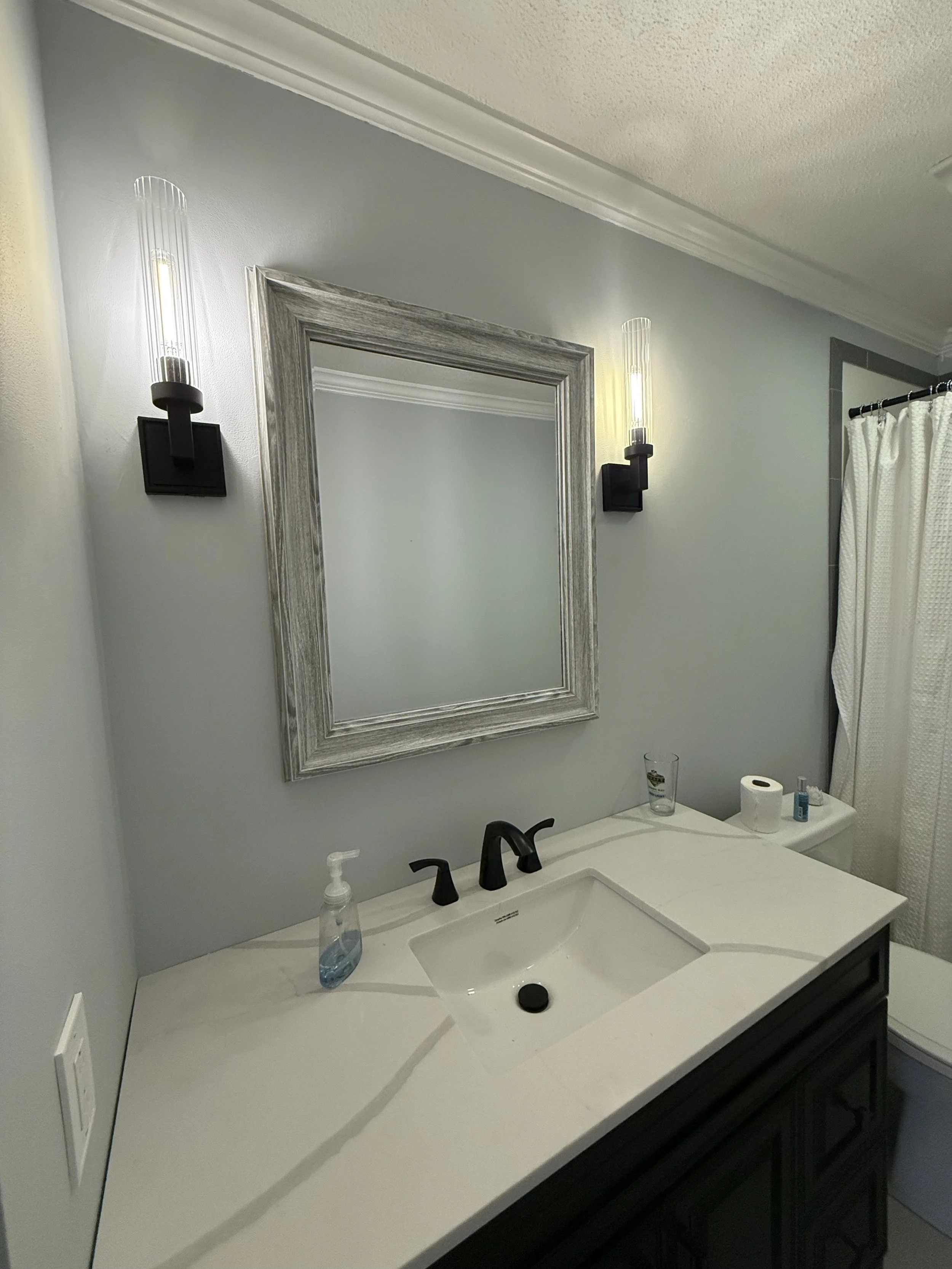 Bathroom vanity with a white countertop, black faucet, mirror framed with gray wood, two modern wall sconces, soap dispenser, glass of water, and toilet paper roll. A shower curtain is partially visible on the right.