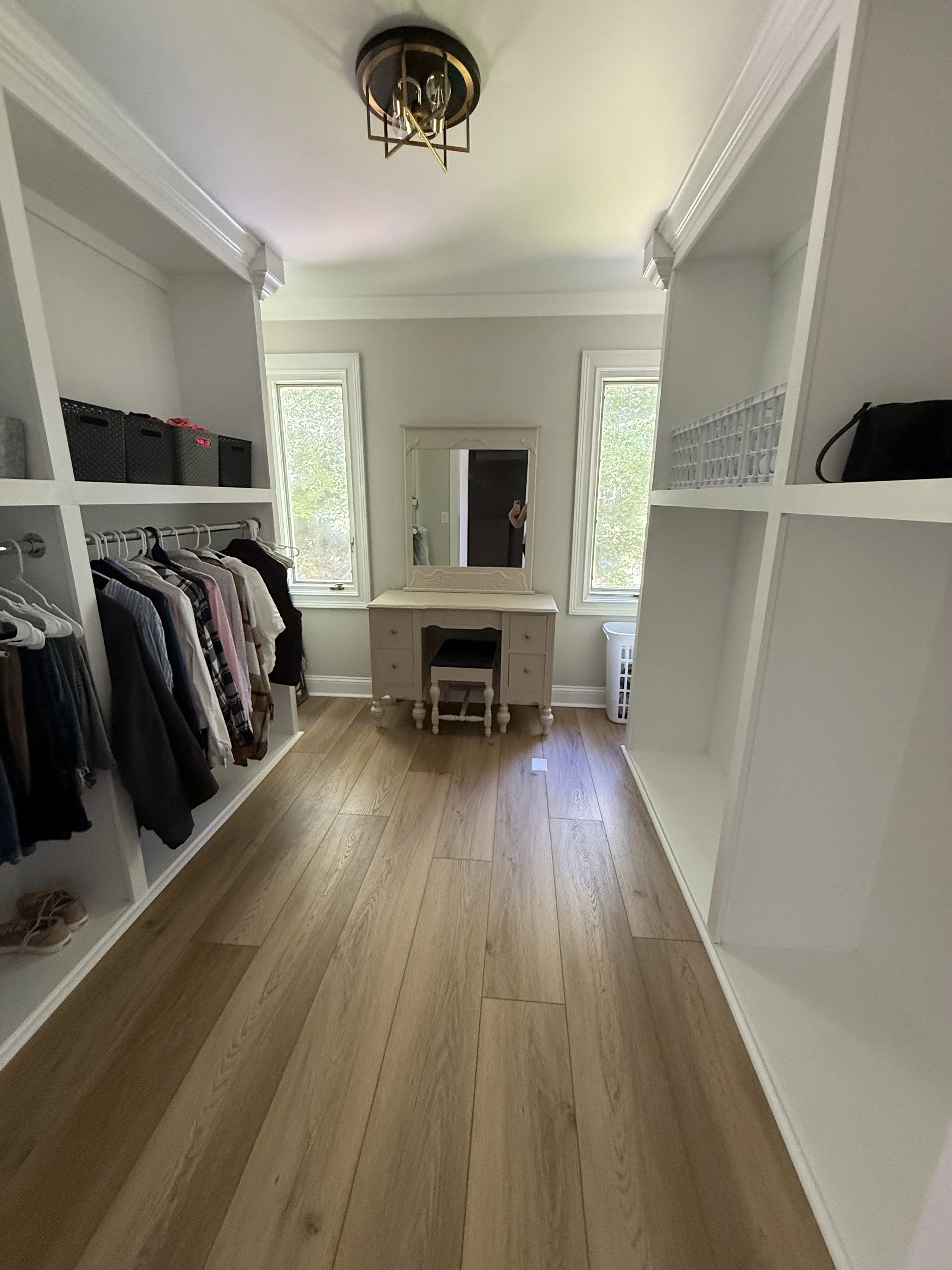 A walk-in closet with white built-in shelves on both sides, clothing hanging on the left, a dresser with a mirror in the center, and two windows letting in natural light.