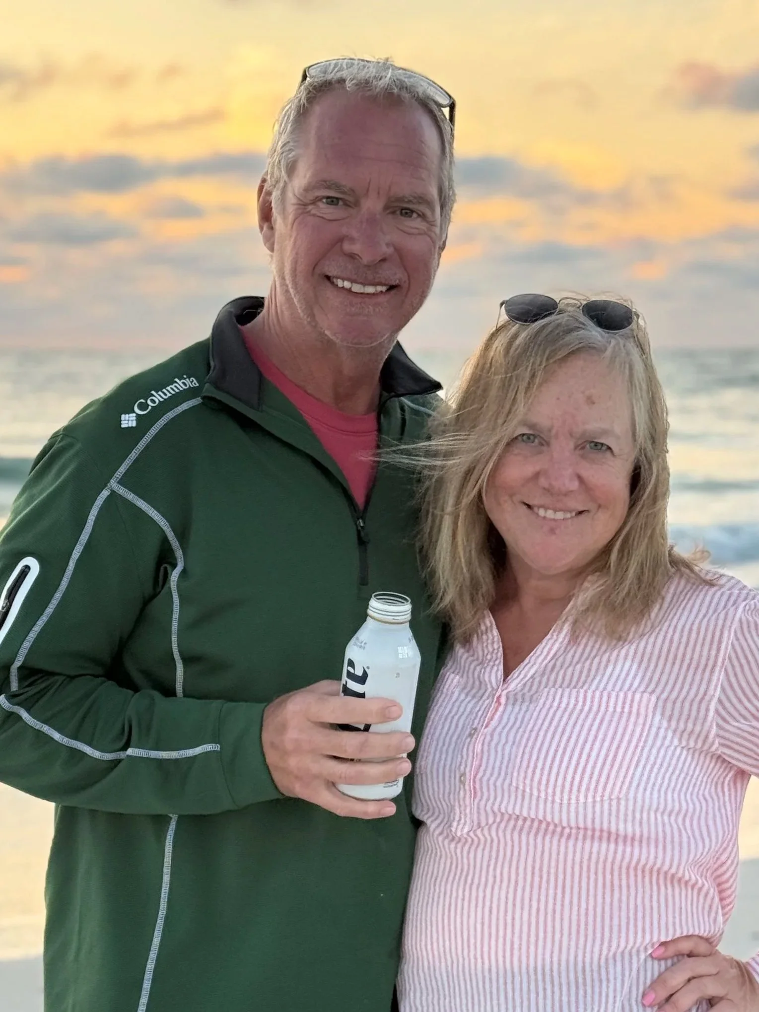 A man and woman standing together on a beach at sunset, smiling. The man is  wearing a green Columbia jacket, the woman is wearing a pink and white striped shirt with sunglasses on her head.