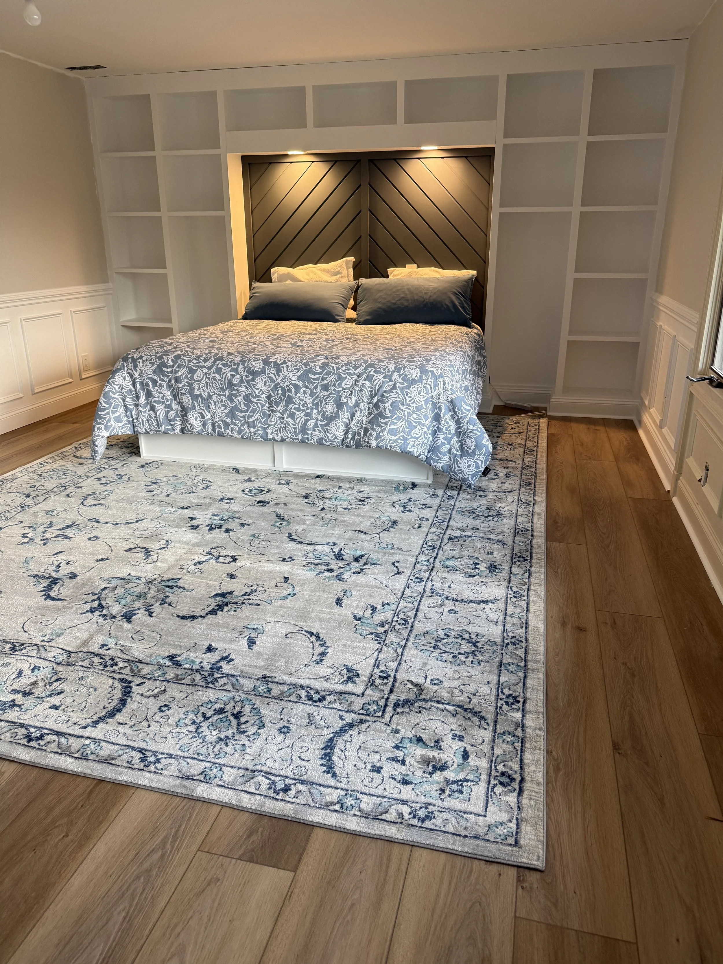 A bedroom with a bed, a patterned blue and white rug, and white oak luxury vinyl plank  floors. The bed has a dark blue wood headboard with diagonal lines, with pillows on top and built-in lighting above.