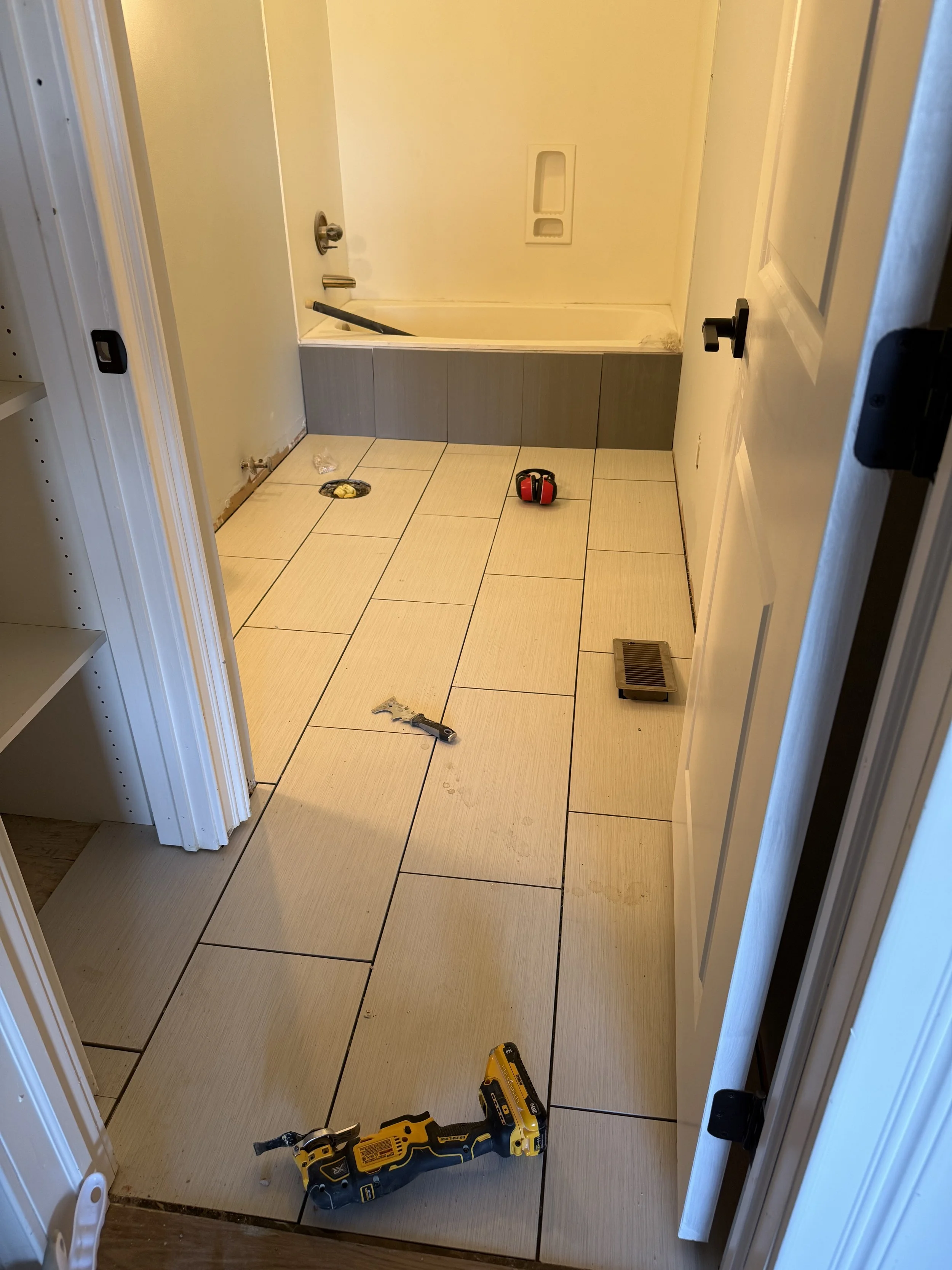 A bathroom under renovation with tools on the tiled floor, including a cordless drill, a wrench, and a yellow tool kit. The bathtub area appears to be partially unfinished.