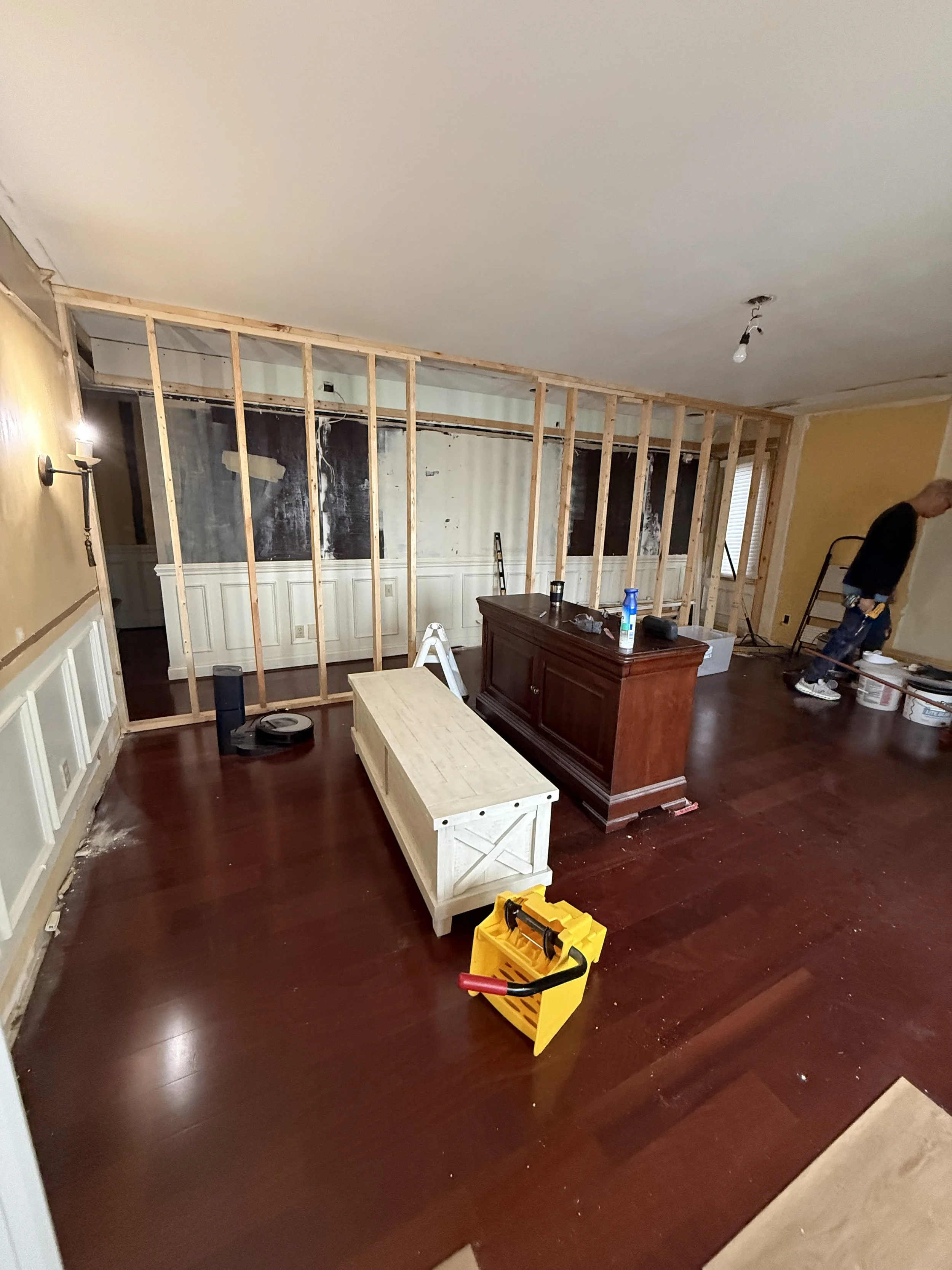Living room undergoing renovation with exposed wall studs, a worker, tools, and furniture.