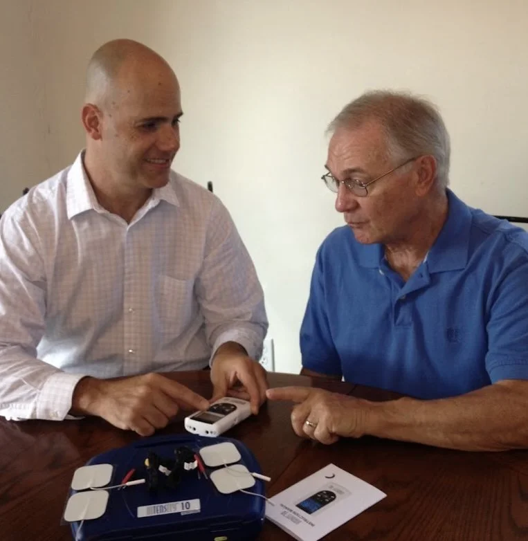 Two men sitting at a table engaging with a medical device, one in a white shirt and the other in a blue polo, with informational papers and a device with wires on the table.