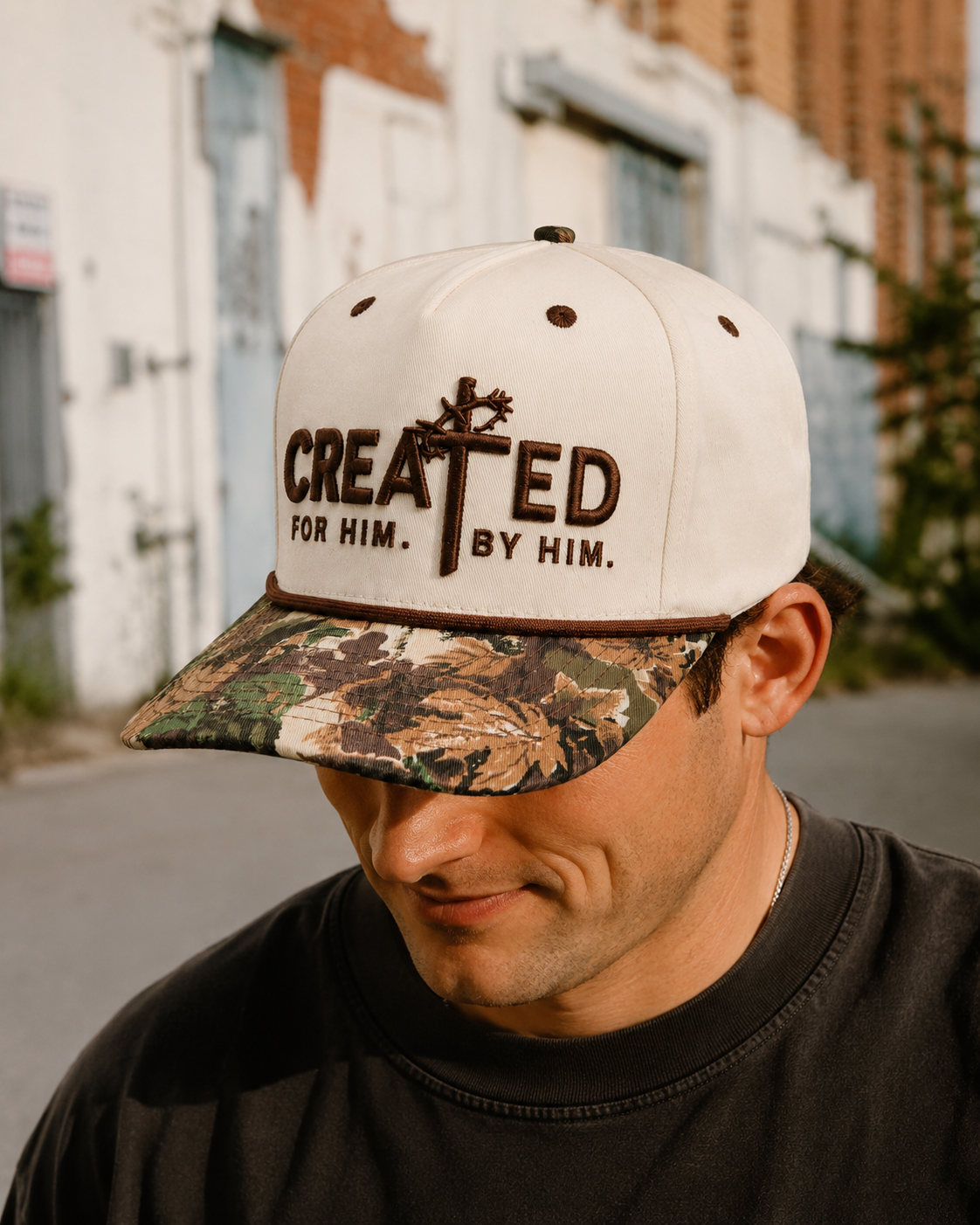 Man wearing a white and camouflage baseball cap with the words 'Created for Him. By Him.' embroidered on the front, standing outdoors with a blurred urban background.