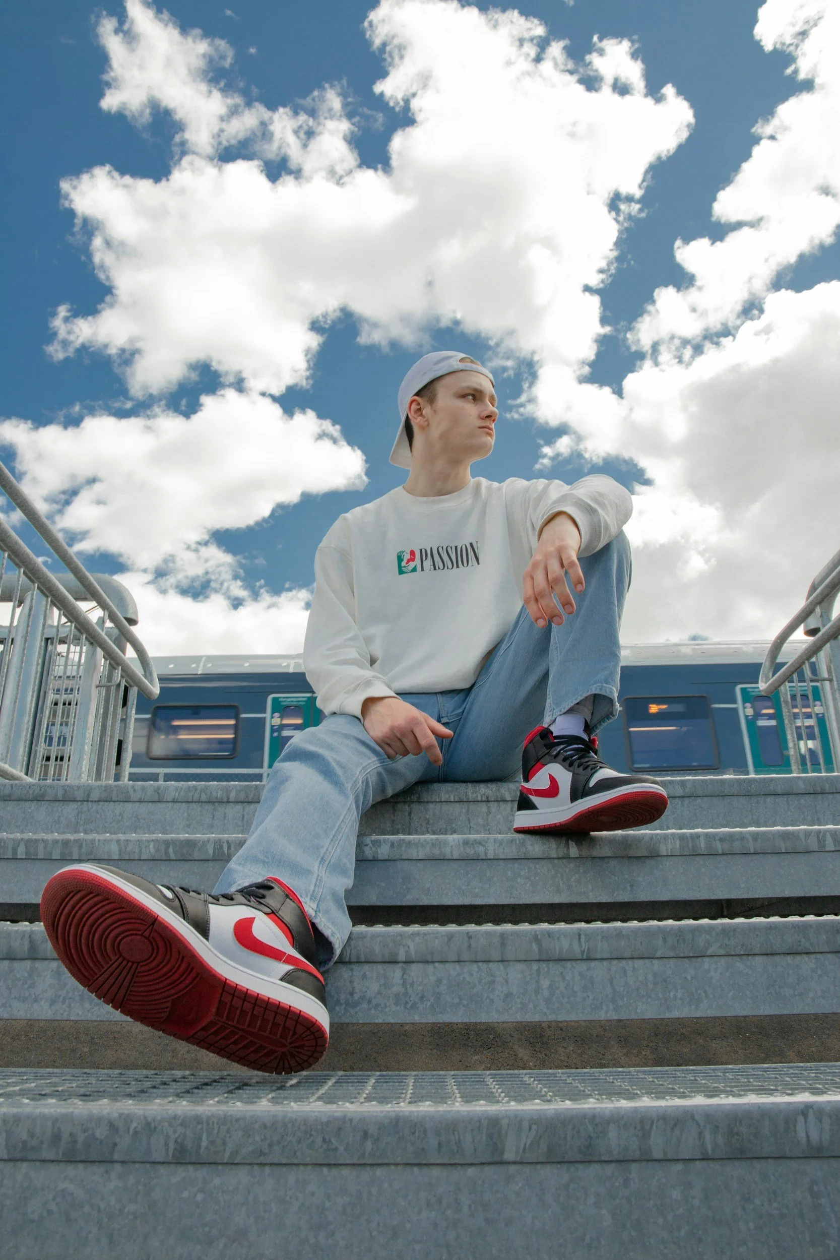 Young man sitting on outdoor stairs under partly cloudy sky, wearing a white sweatshirt with 'Passion' logo, white cap backward, blue jeans, and red, white, and black sneakers.