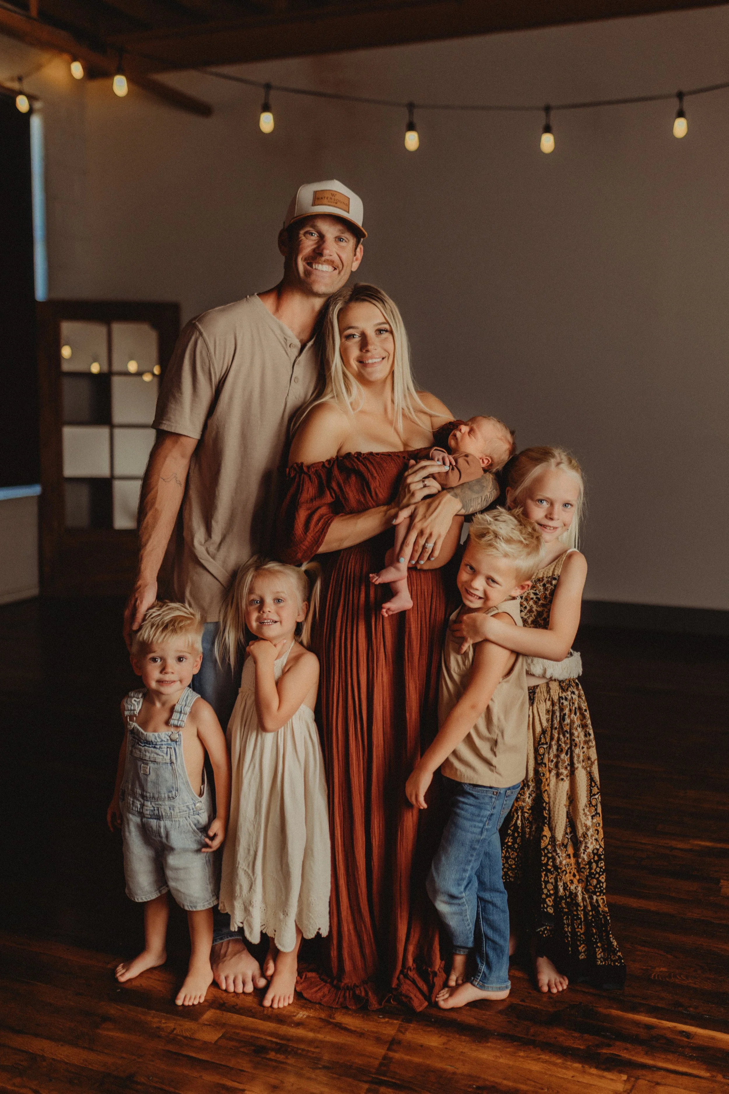 Family of six posing for a photo indoors under string lights, with one woman holding a baby, four children, and a man standing behind them. All are smiling, and the atmosphere is warm and cozy.