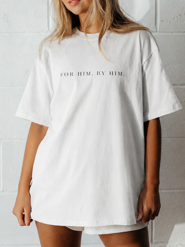 Woman wearing a white T-shirt with the text 'For Him. By Him.' printed on it, standing against a light-colored brick wall.