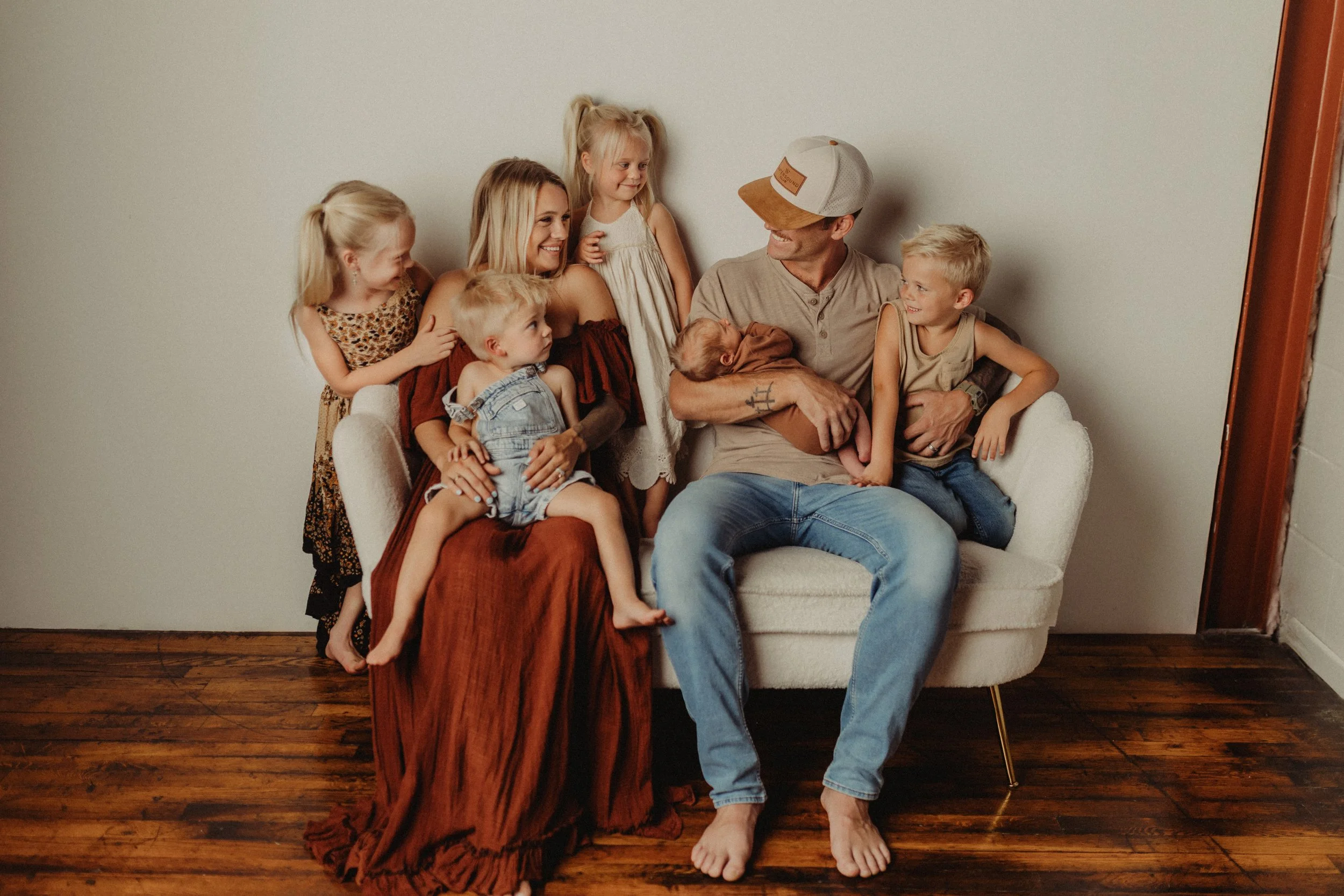 A family sitting on a white couch, smiling and looking at each other. The family includes an adult woman, an adult man wearing a cap, and five children around them. Some children are sitting on the couch, while others are standing or leaning on it. The background is plain with a wooden floor visible.