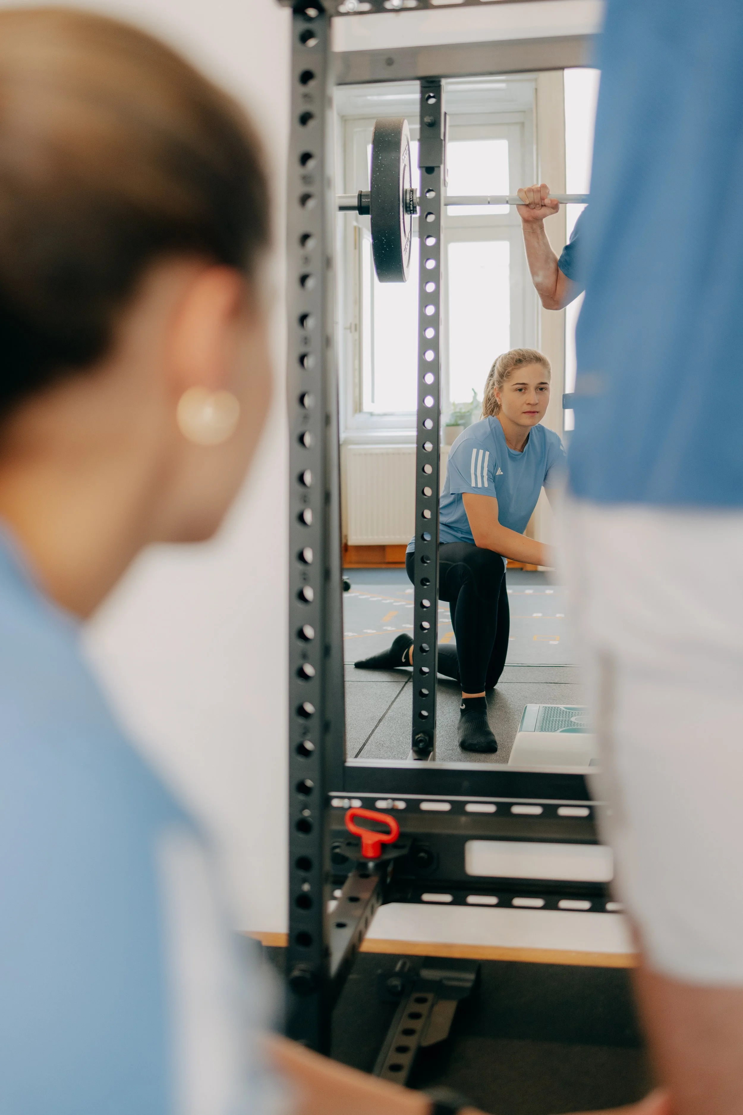 Ein Mann hebt eine Langhantel beim Krafttraining, während eine junge Frau in sportlicher Kleidung auf dem Boden kniet und zuschaut, in einem Fitnessraum mit großen Fenstern.