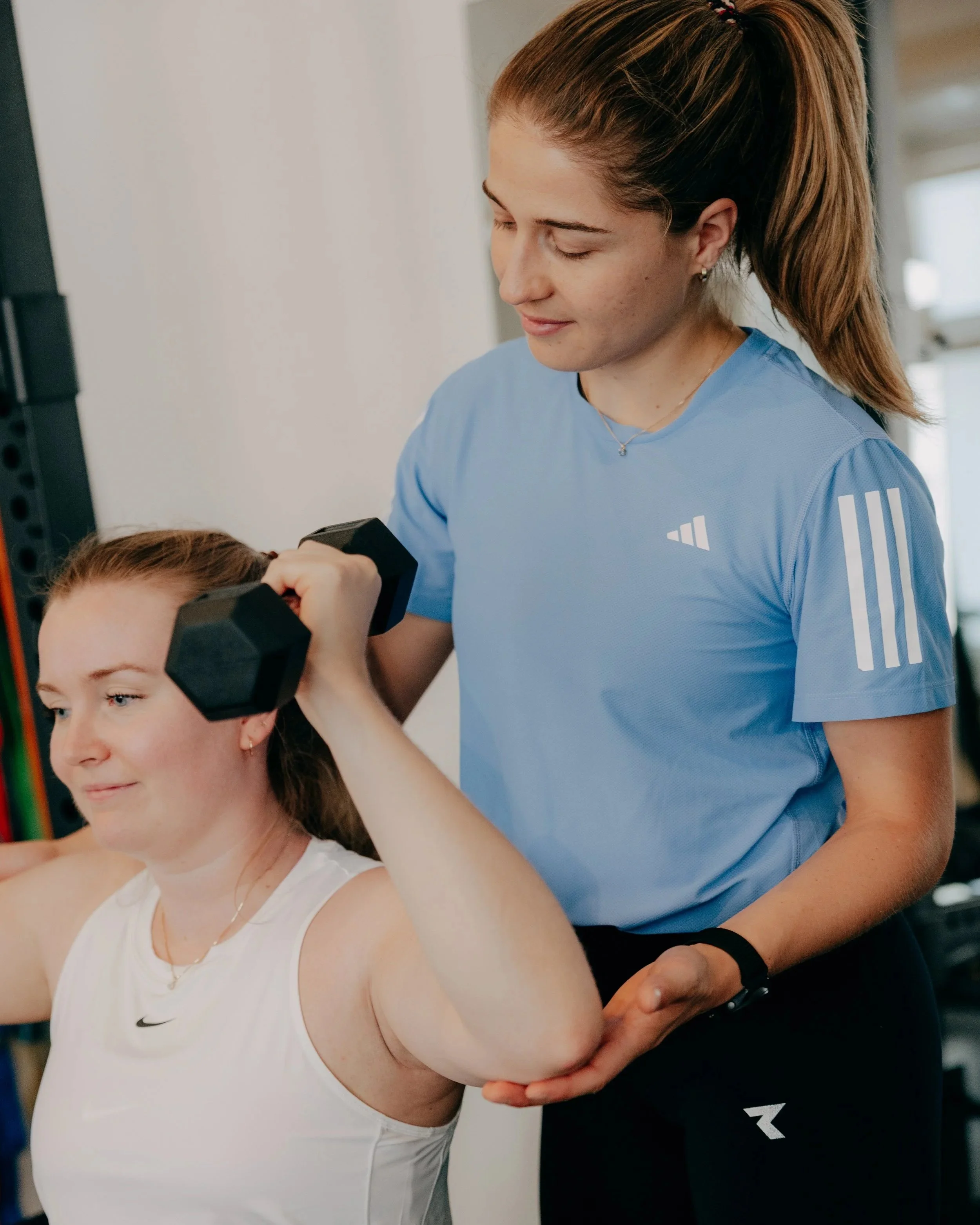 Eine junge Frau beim Krafttraining, während eine Trainerin unterstützend ihre Muskulatur behandelt.