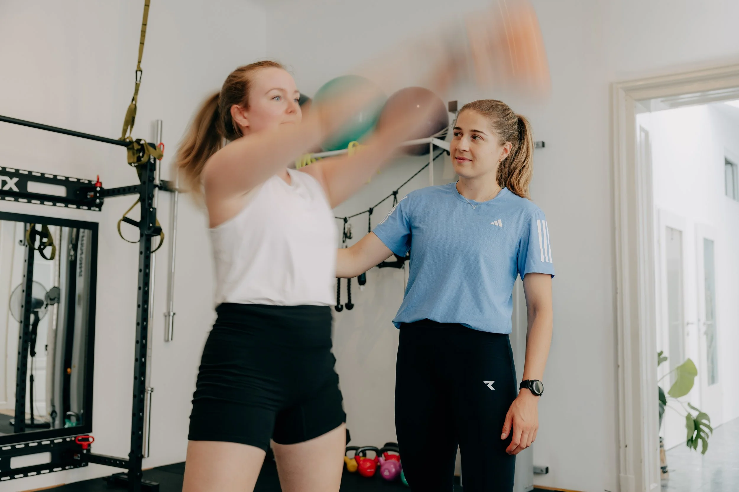 Zwei junge Frauen trainieren im Fitnessstudio mit einer Gymnastikball und einer Trainerin, die sie bei ihrer Übung unterstützt.