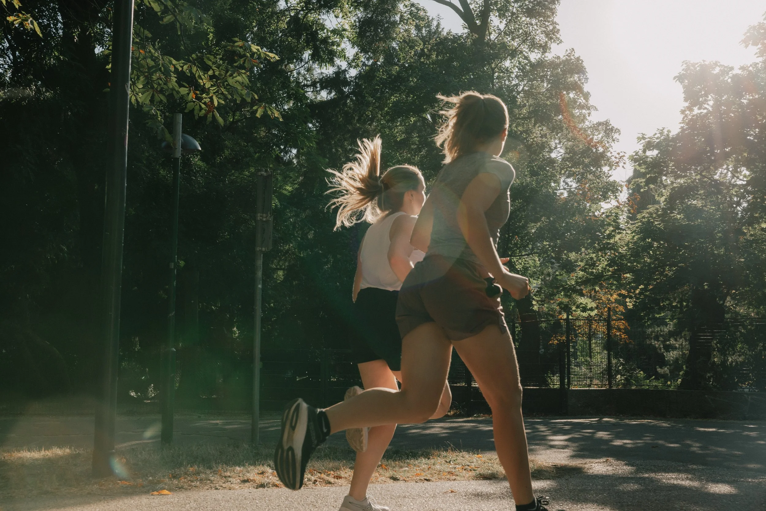 Zwei junge Frauen laufen in einem Park an einem sonnigen Tag, umgeben von Bäumen.