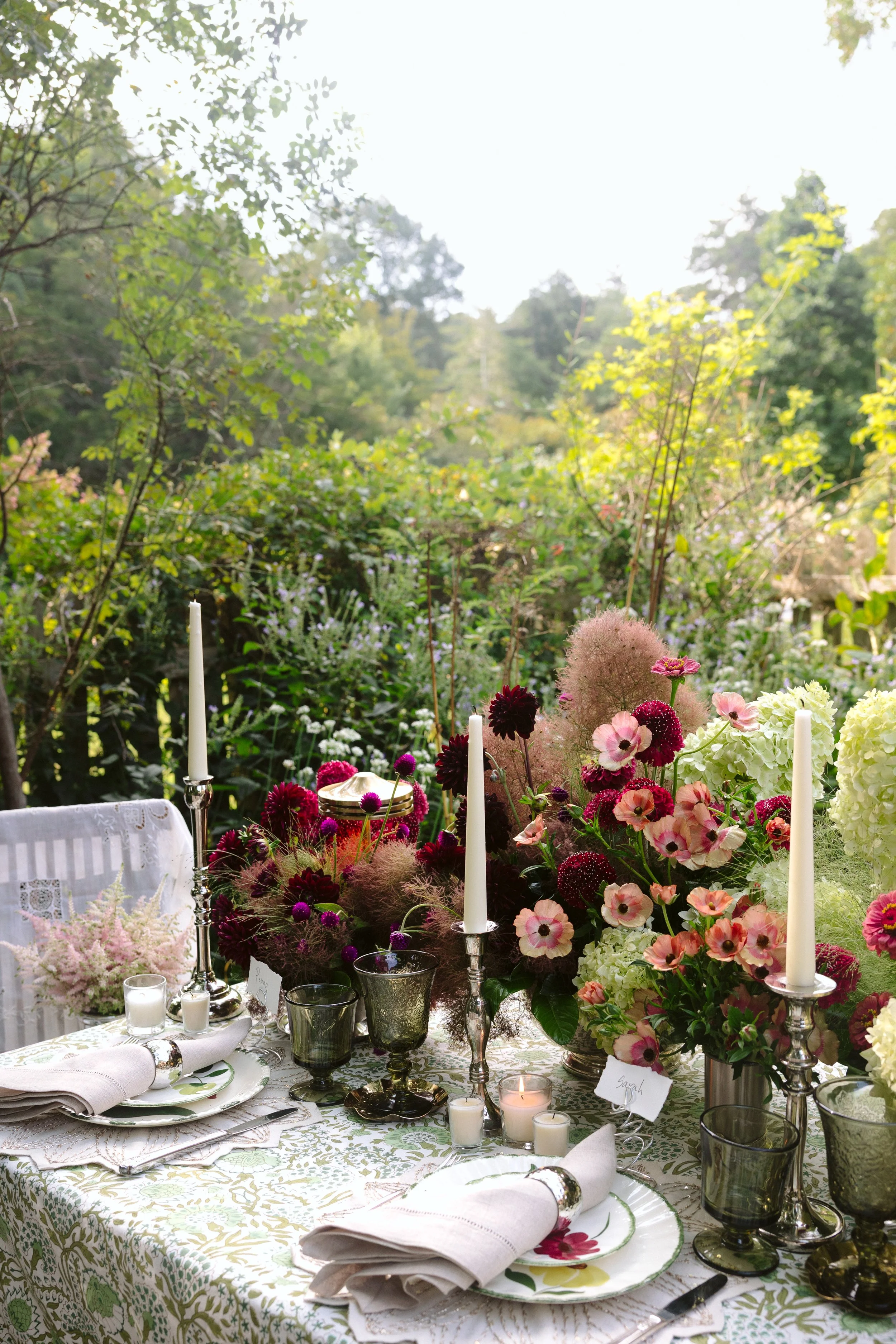 A beautifully decorated outdoor dining table with floral arrangements, candles, and fine china, set in a lush garden with trees and greenery in the background.
