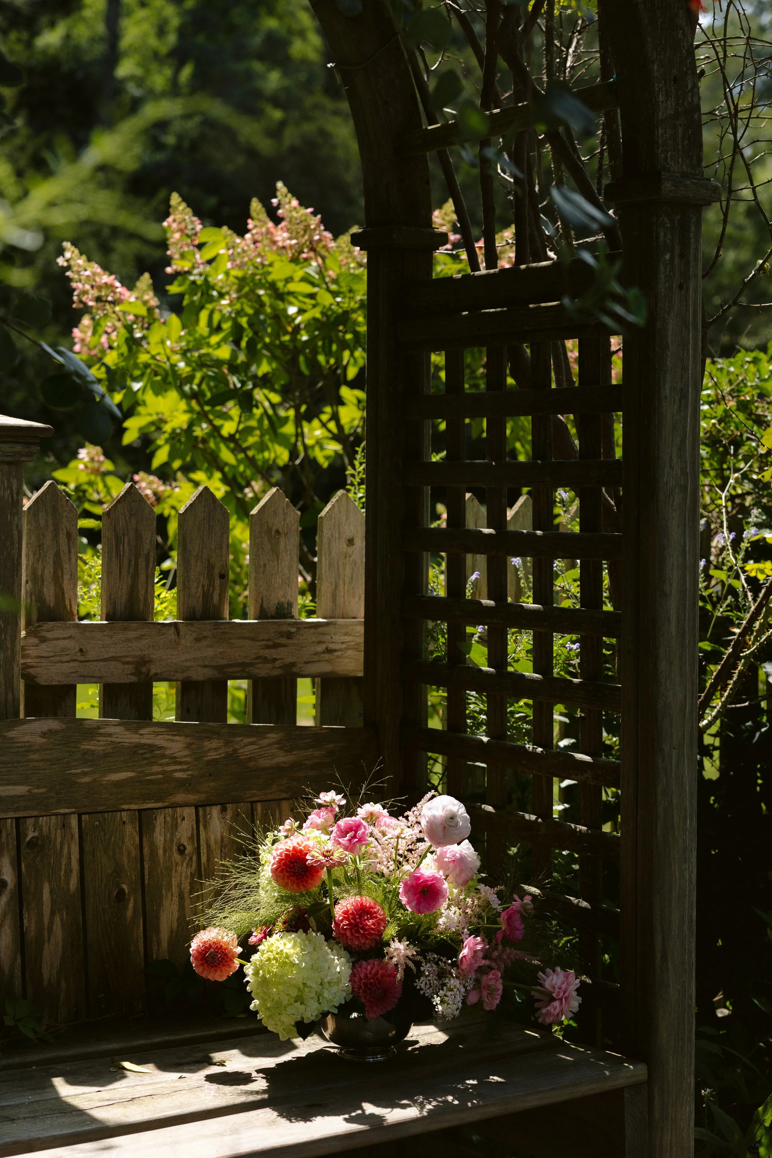 Brightly lit garden scene with a wooden picket fence, a wooden gate, and a flower arrangement in a black vase featuring pink, white, and green blossoms.