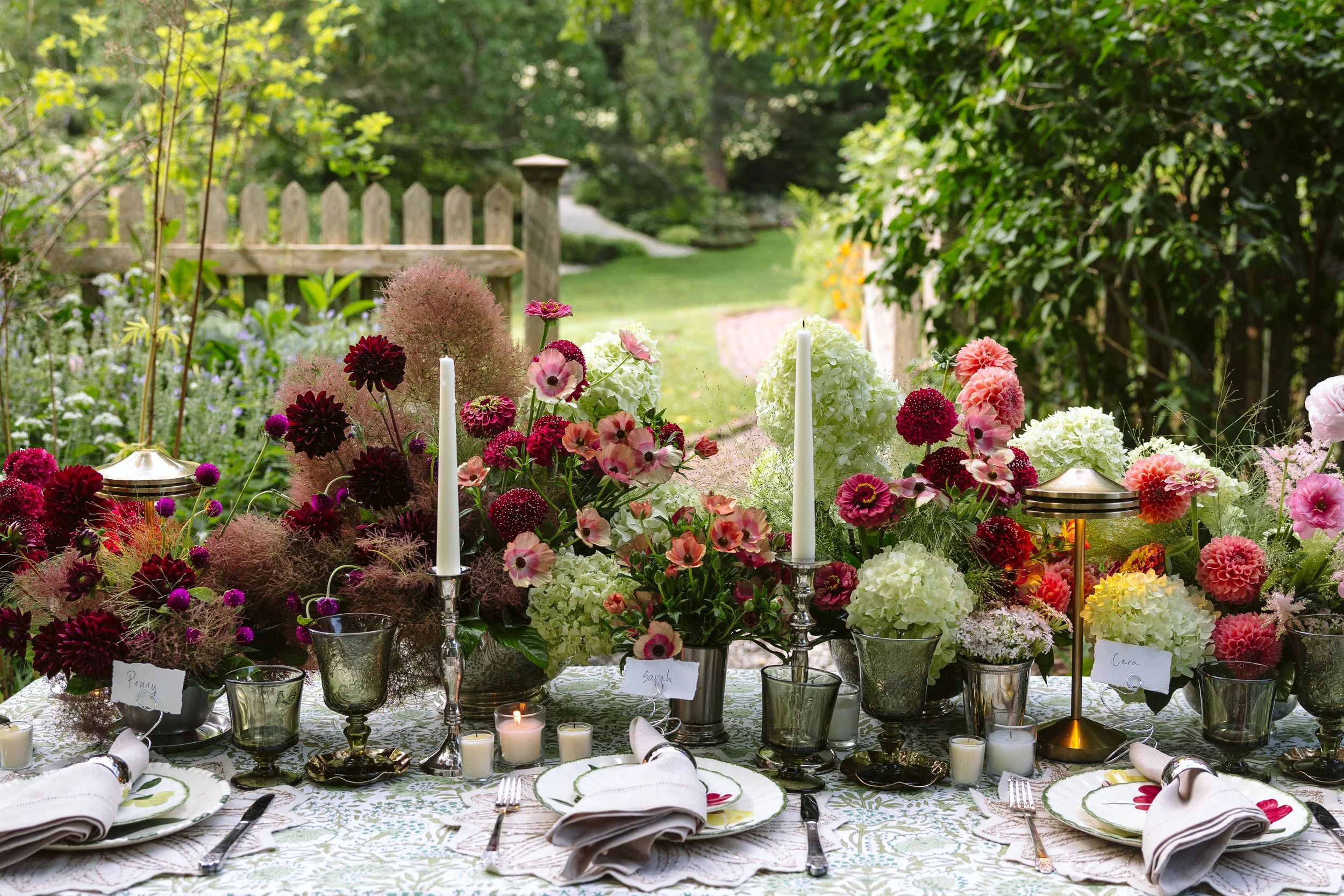 A decorated outdoor dining table with floral arrangements, candles, and place settings, set in a lush green garden.