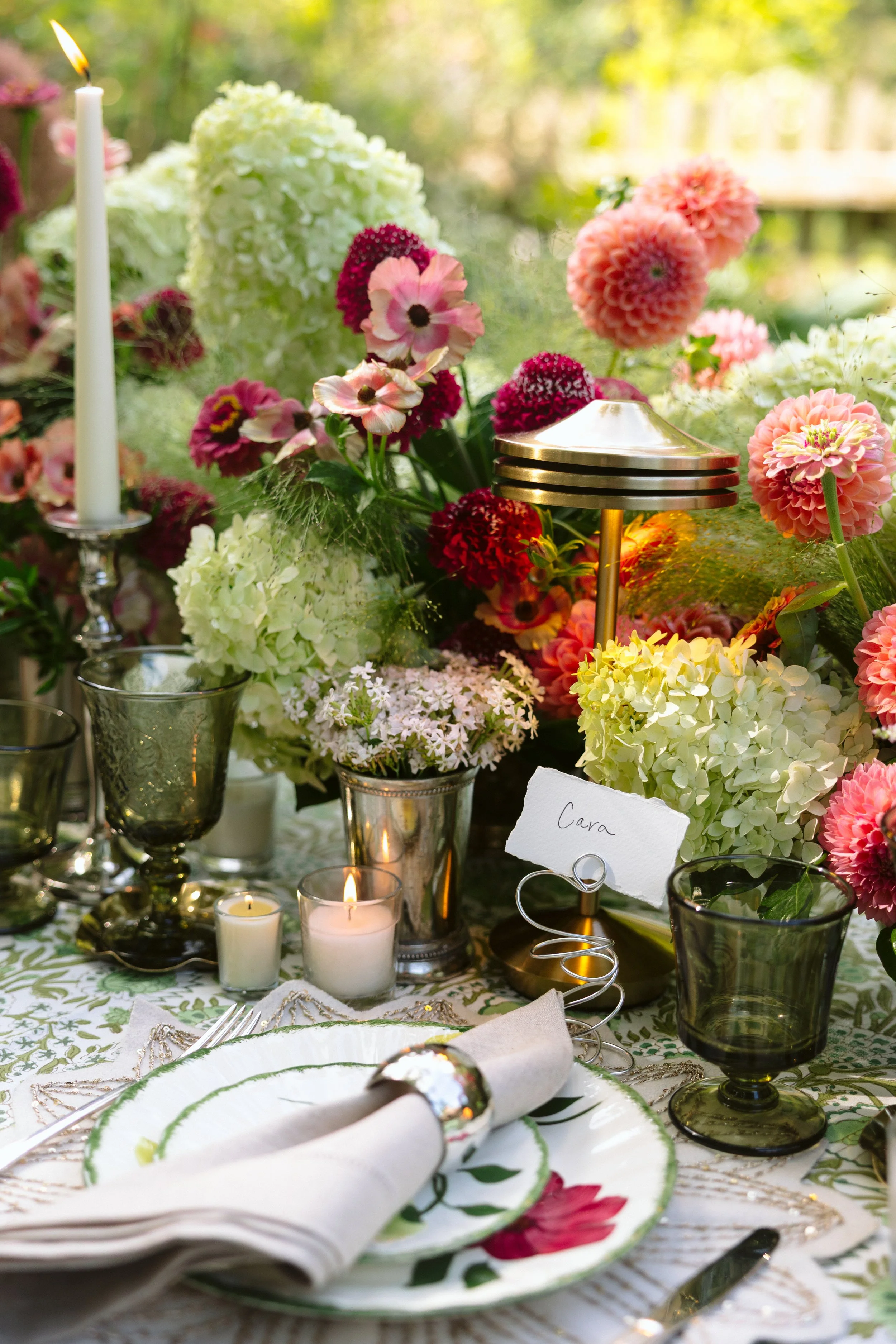 A beautifully decorated table with floral arrangements, candles, and fine china, set outdoors in a garden or park.