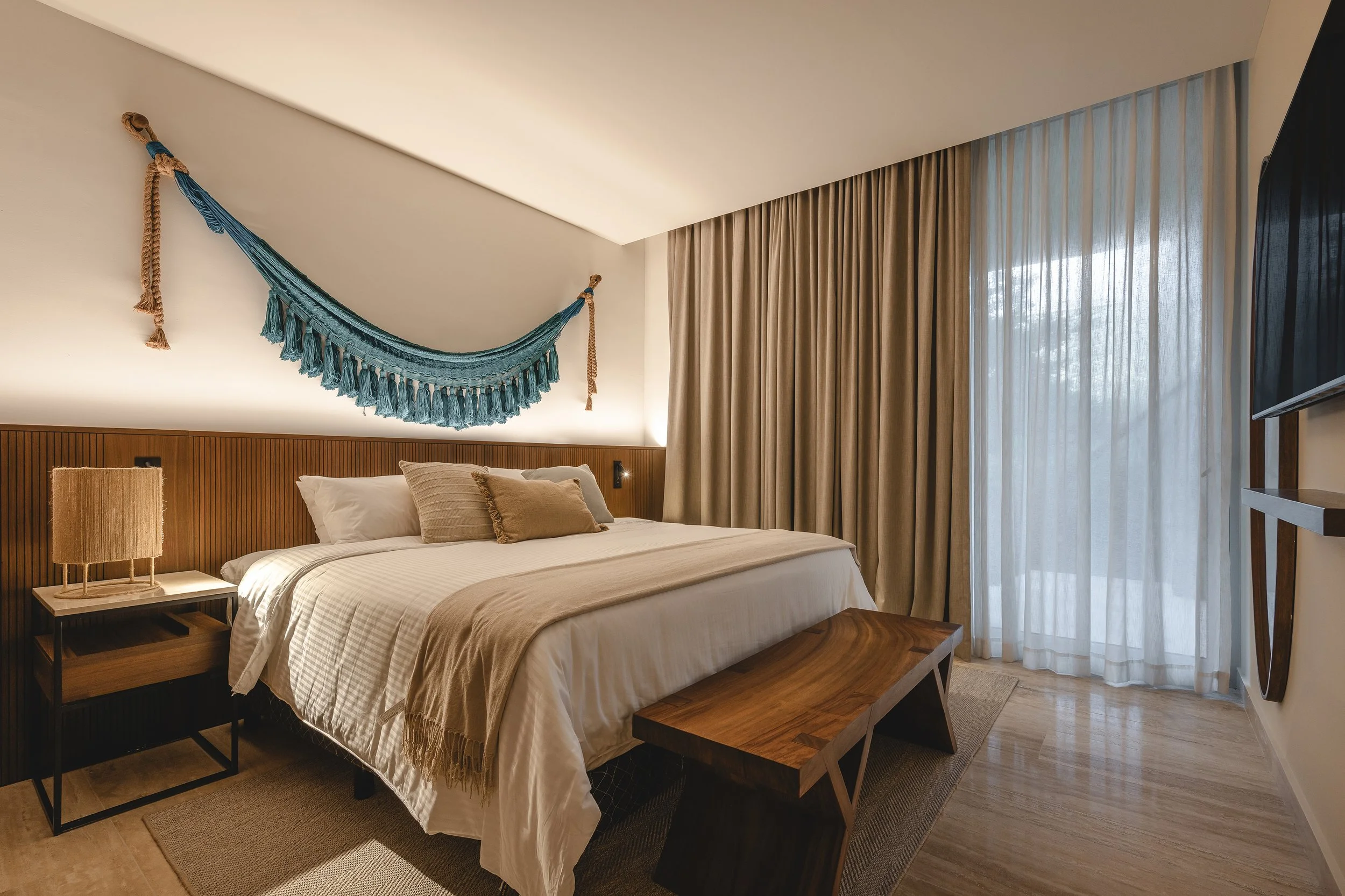 Habitación moderna con cama doble, cojines, lámpara de mesa, cortinas largas y televisión en la pared.