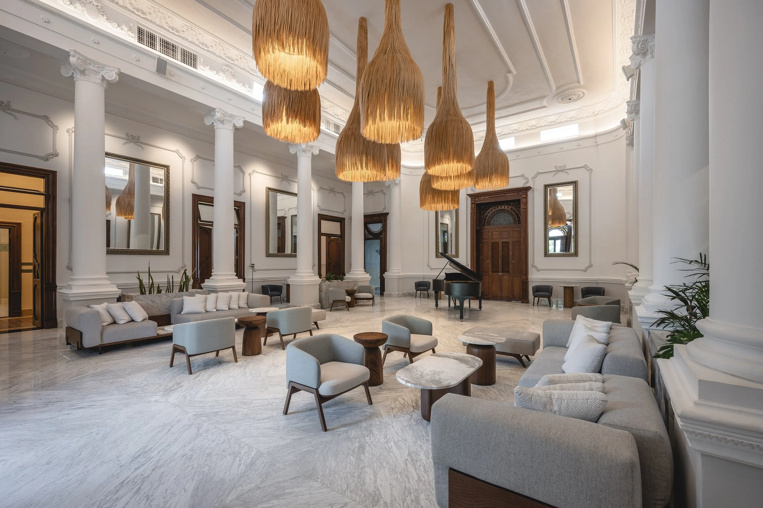 Vestíbulo o sala de estar con columnas blancas y detalles decorativos en el techo, muebles de sala en tonos neutros, un piano de cola negro, espejos en las paredes y grandes lámparas decorativas colgando del techo de fibra natural.