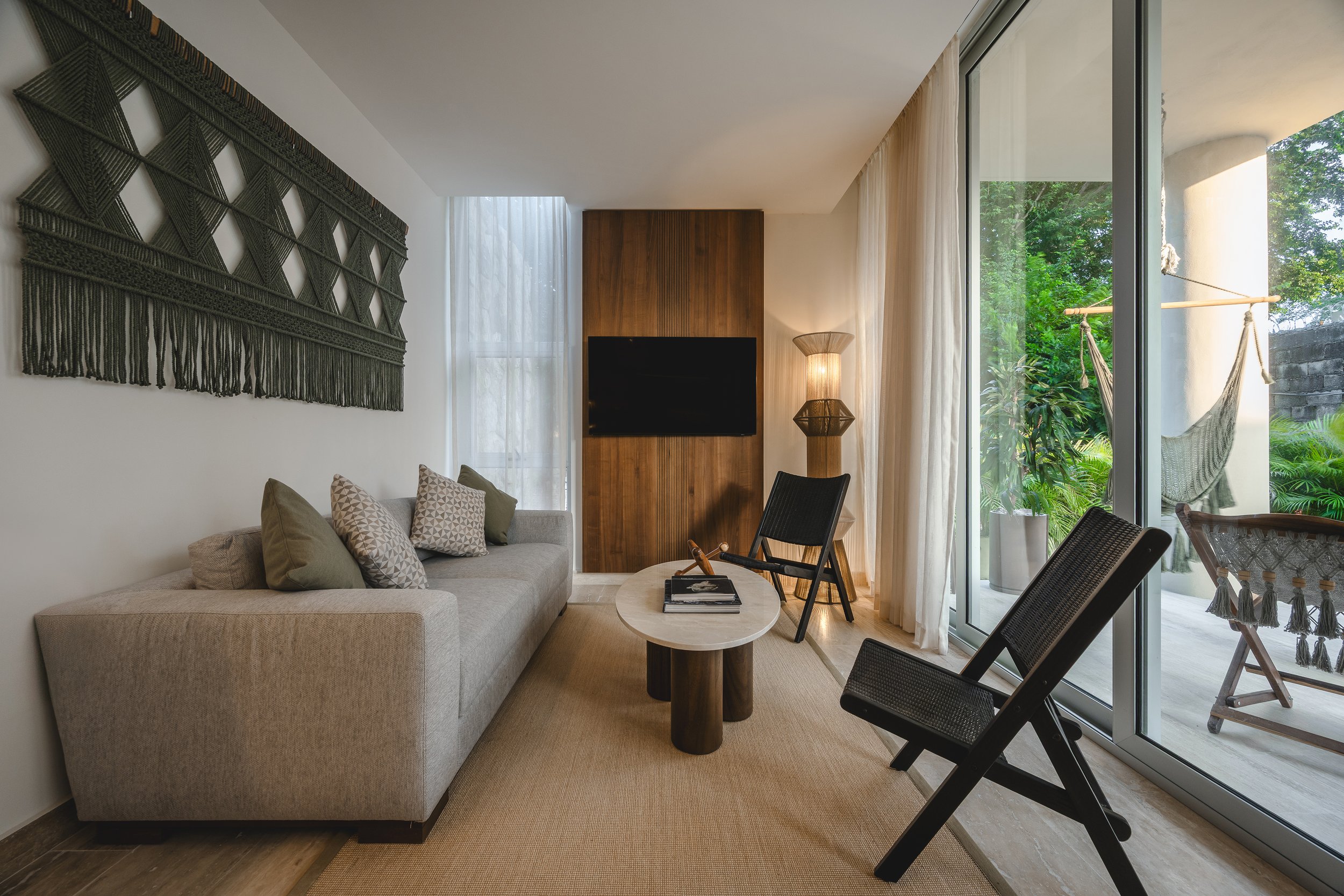 Sala de estar con sofá beige, mesa redonda y sillas negras, paredes blancas con arte textil y una pared de madera con televisor, ventanas grandes con cortinas claras y vista a un balcón con hamaca y plantas verdes.