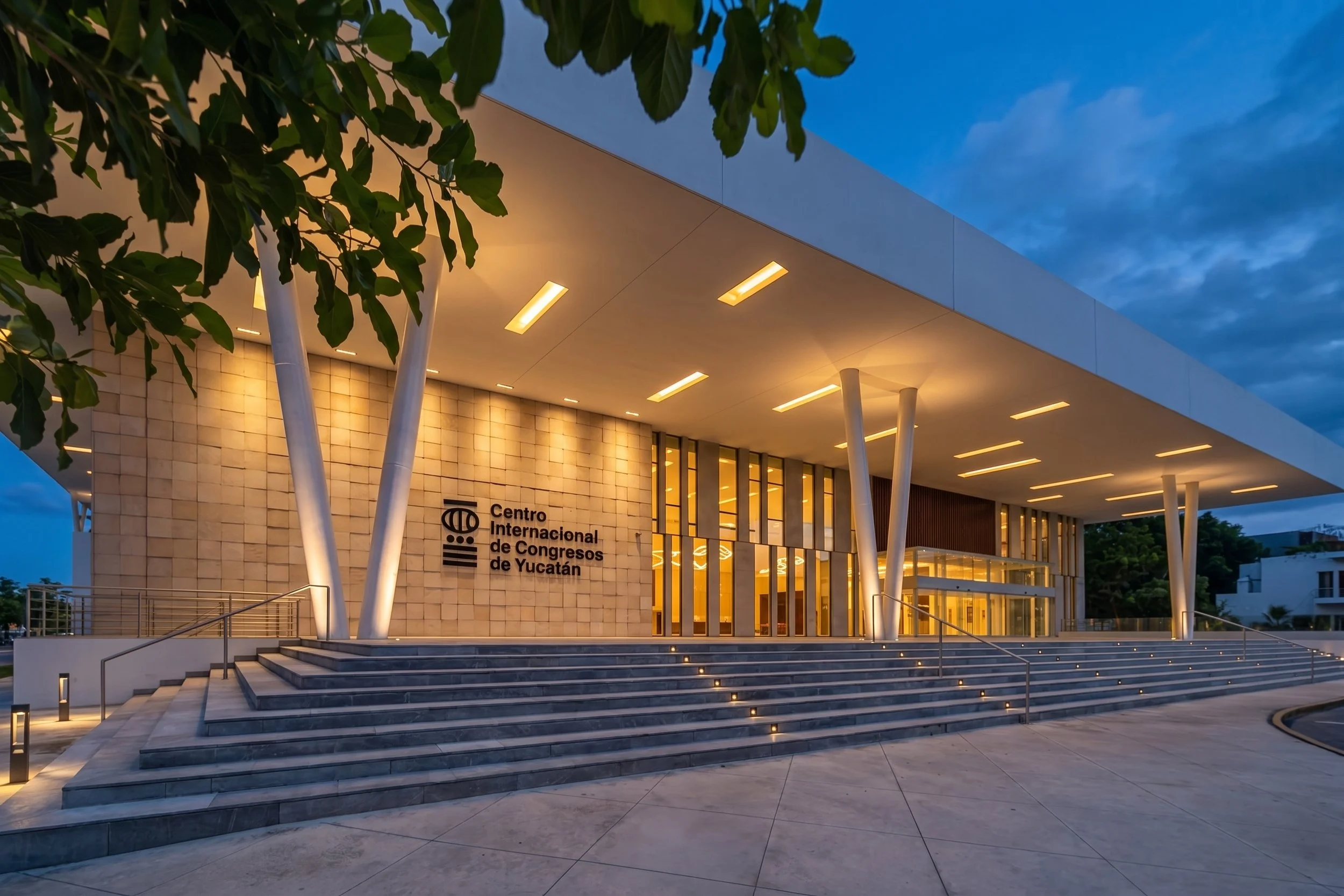 Edificio del Centro Internacional de Congresos de Yucatán visto en la noche, con luces encendidas, escalones y jardines delanteros, rodeado de árboles y cielo despejado con nubes.