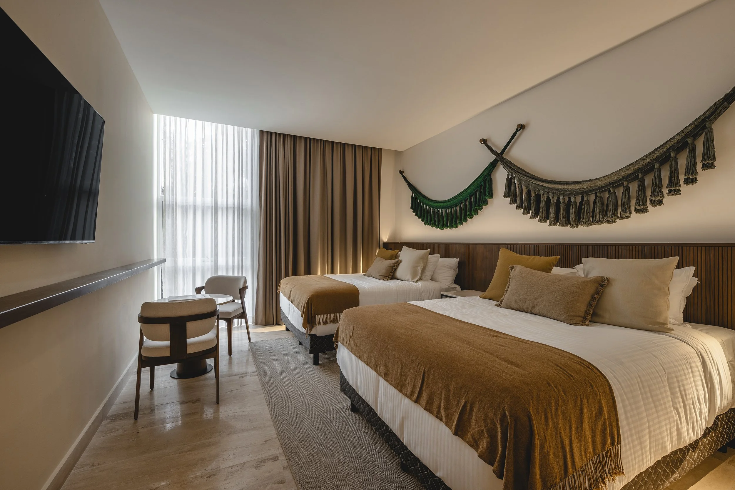 Habitación de hotel con dos camas, cortinas largas, una televisión en la pared y decoración con tejidos colgados en la pared.