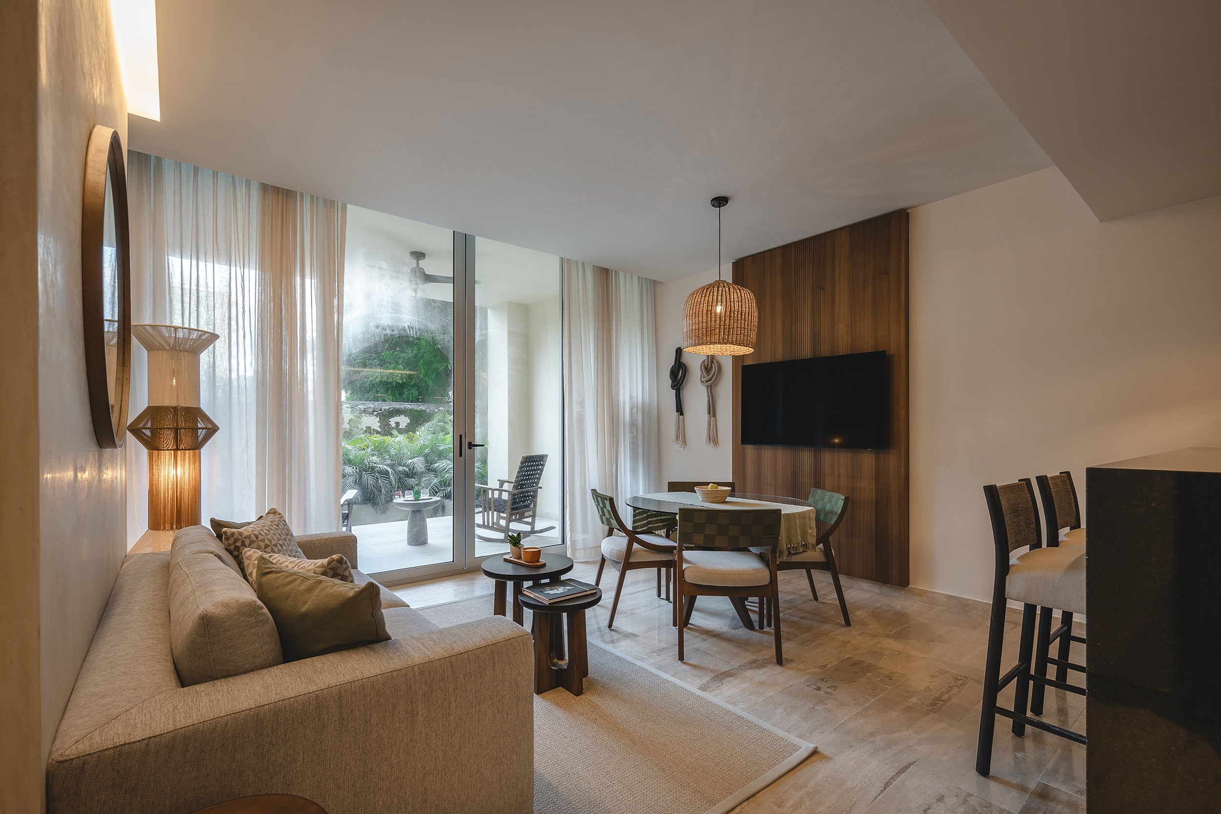 Sala de estar con sofá beige, mesa de centro, sillones y una mesa redonda con plantas, con grandes ventanas con cortinas y una vista a un jardín exterior, decorada con lámparas de fibras naturales y obra de arte en la pared de madera.