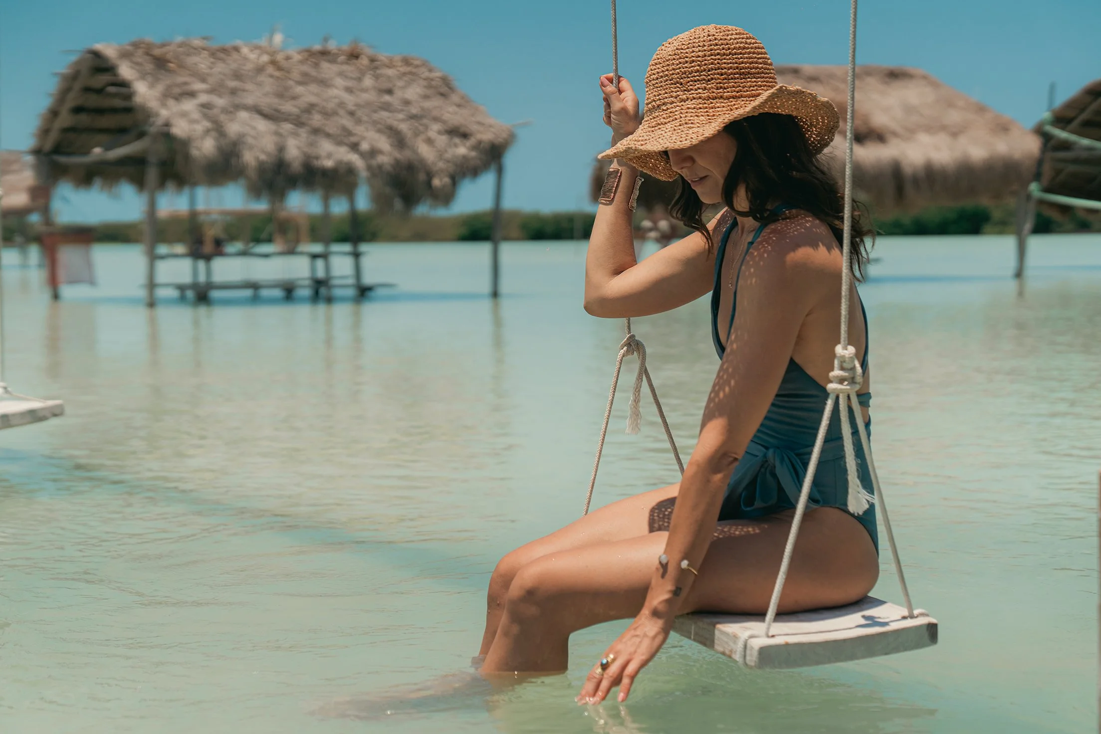 Mujer con sombrero de paja en una hamaca columpio en un entorno de playa con agua cristalina y cabañas de madera con techos de paja.