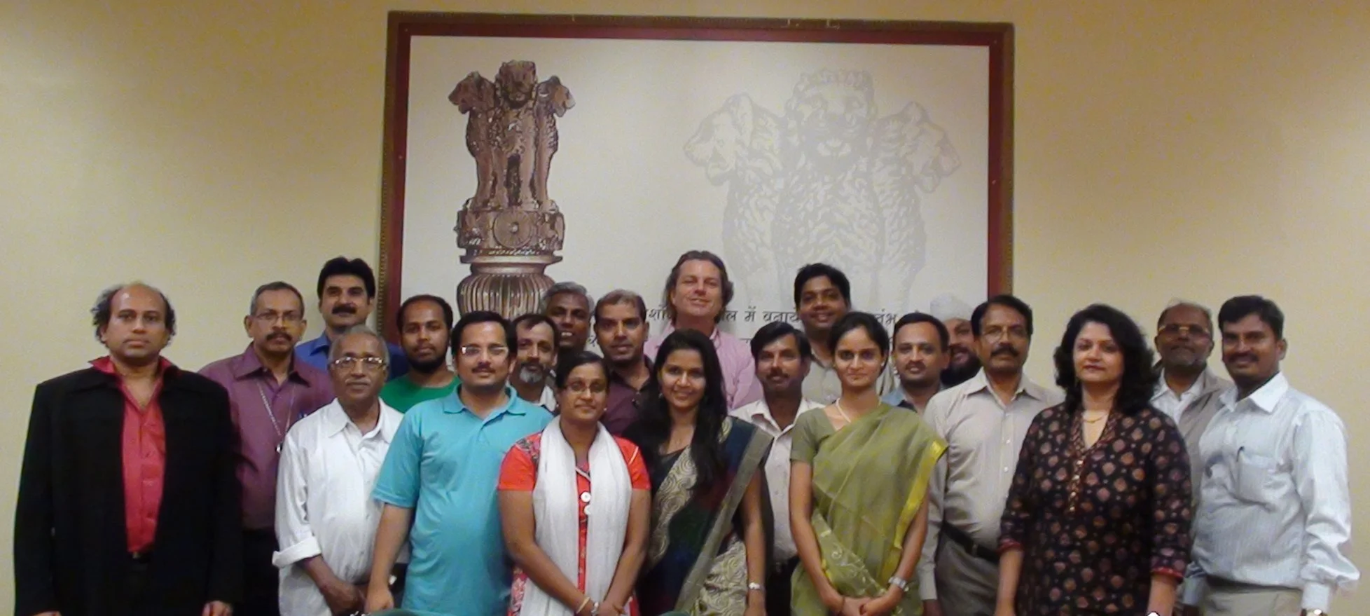 Group of men and women standing together for a photo in front of a wall with an Indian emblem and Hindi script.